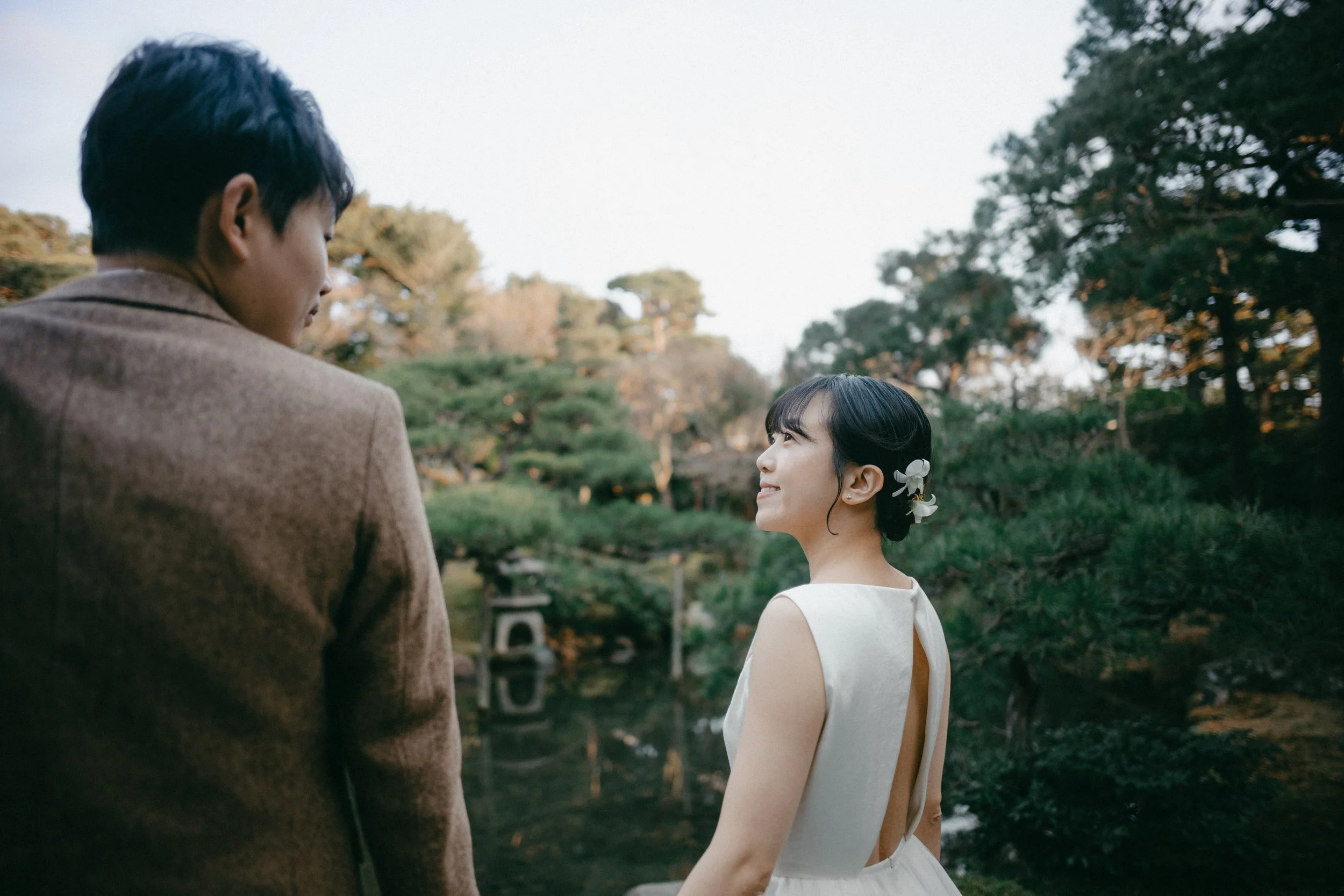 A couple dressed in formal attire standing outdoors near a pond with trees in the background. The woman is smiling and looking at the man, who is facing away from the camera.
