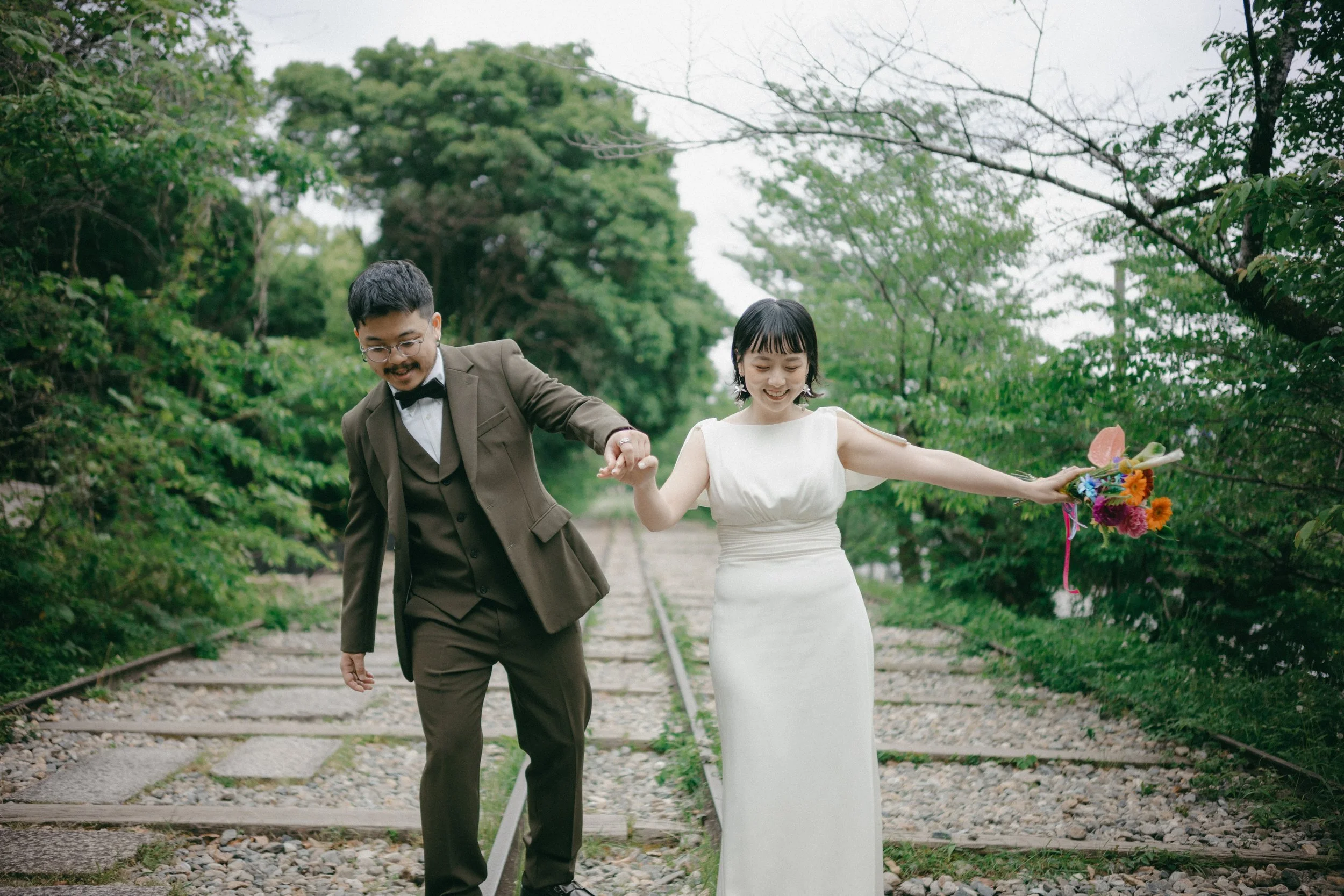 A happily smiling bride and groom holding hands while walking on railway tracks surrounded by greenery. The bride is in a white dress holding a bouquet of colorful flowers, and the groom is in a brown suit with glasses.