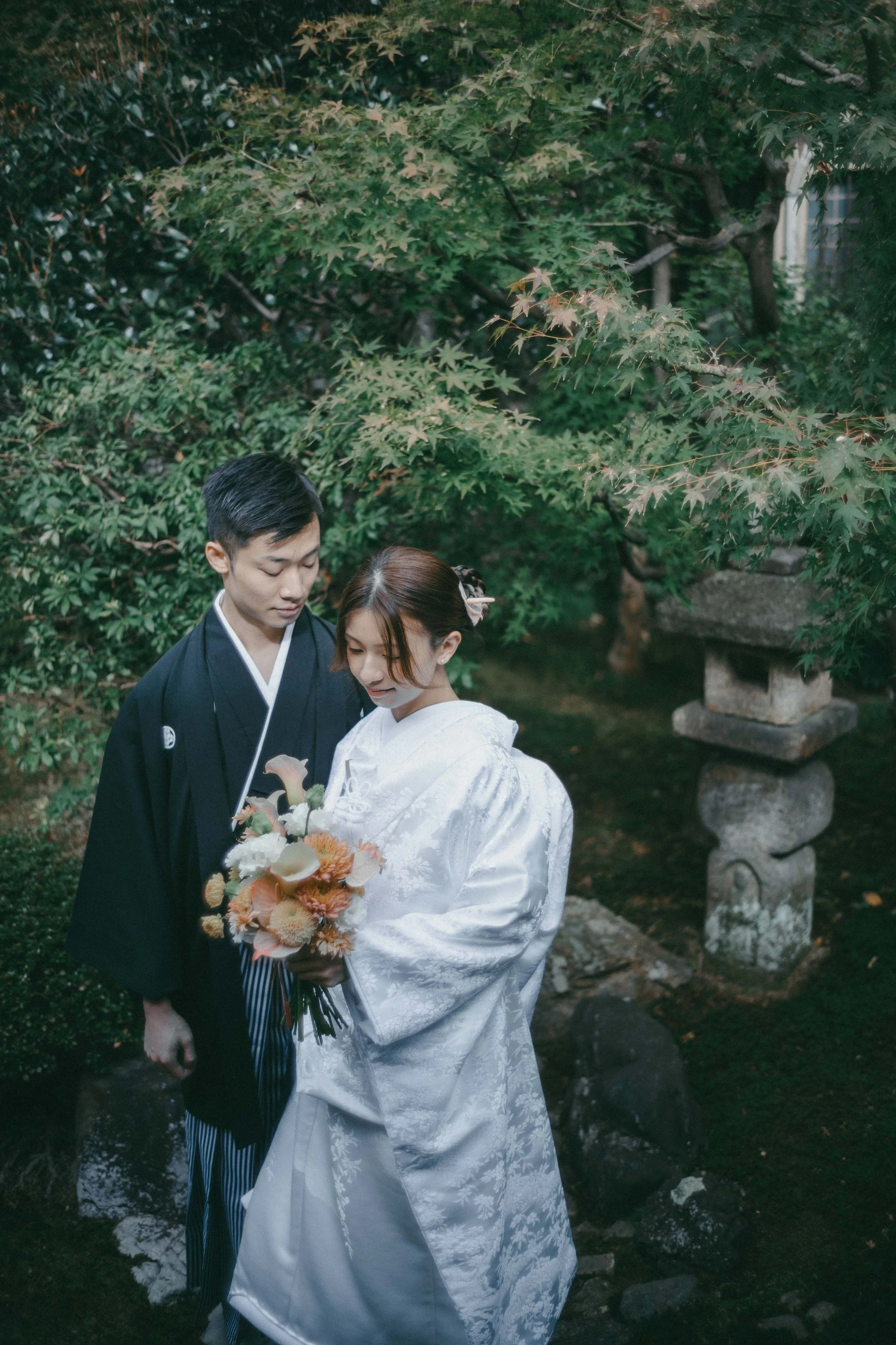 A Japanese bride and groom in traditional attire standing close together outdoors, with greenery and a stone lantern in the background, the bride holding a bouquet of flowers.