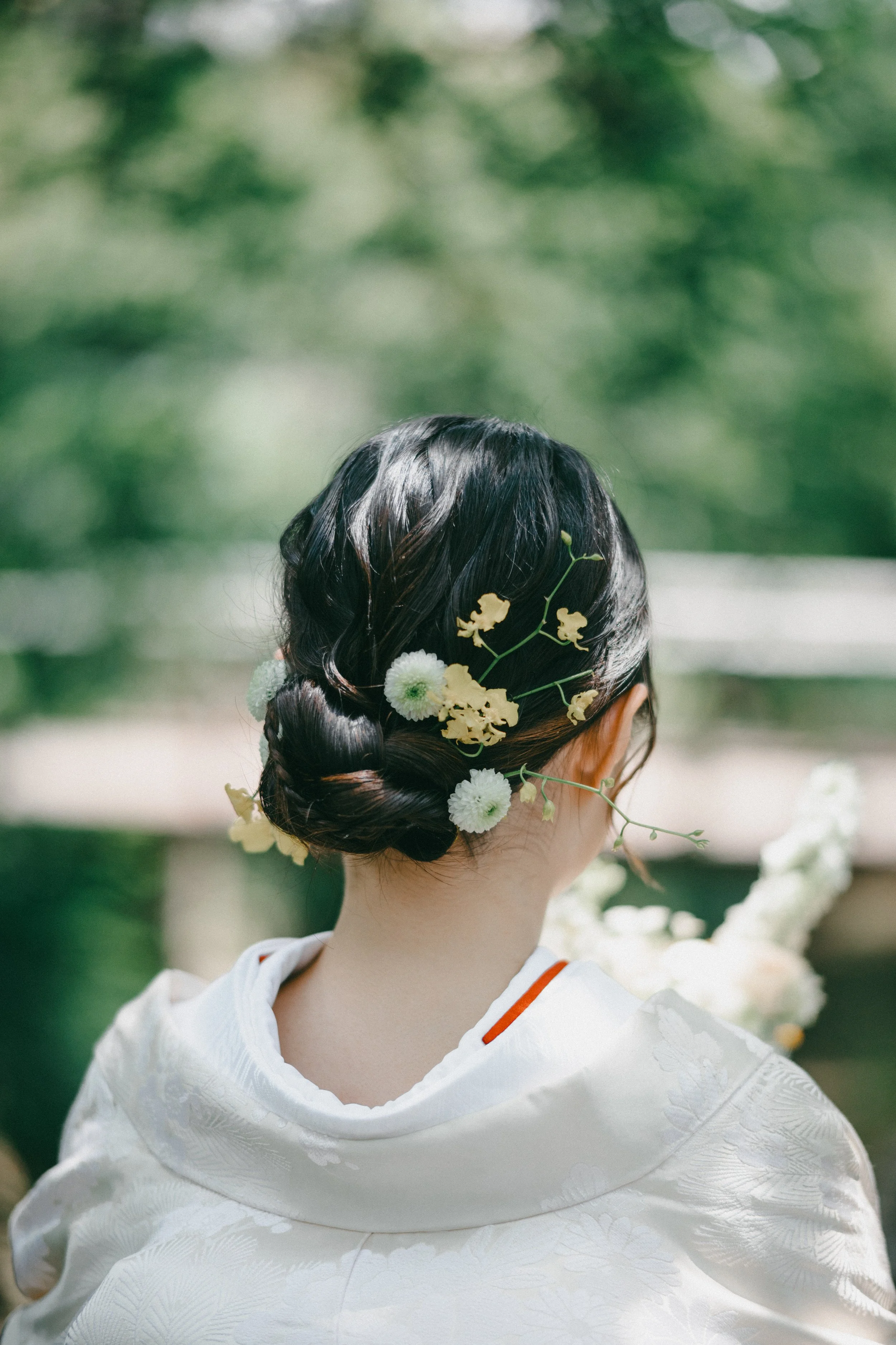 Rear view of a woman with dark hair styled in an elegant updo, adorned with white and yellow flowers, wearing a white traditional kimono, standing outdoors with green foliage in the background.