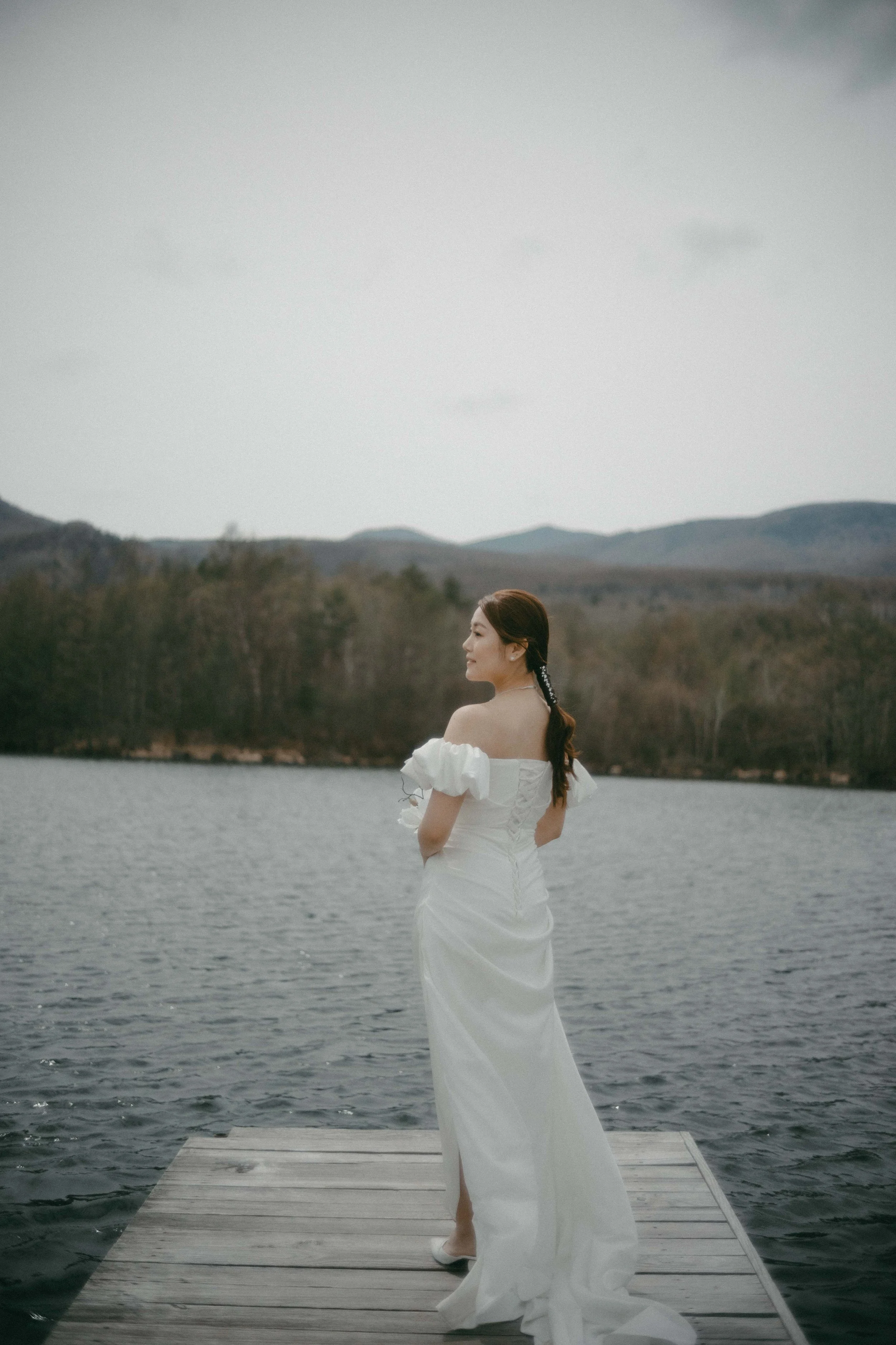 A woman in a white wedding dress stands on a wooden dock by a lake, gazing to the side with mountains and trees in the background.