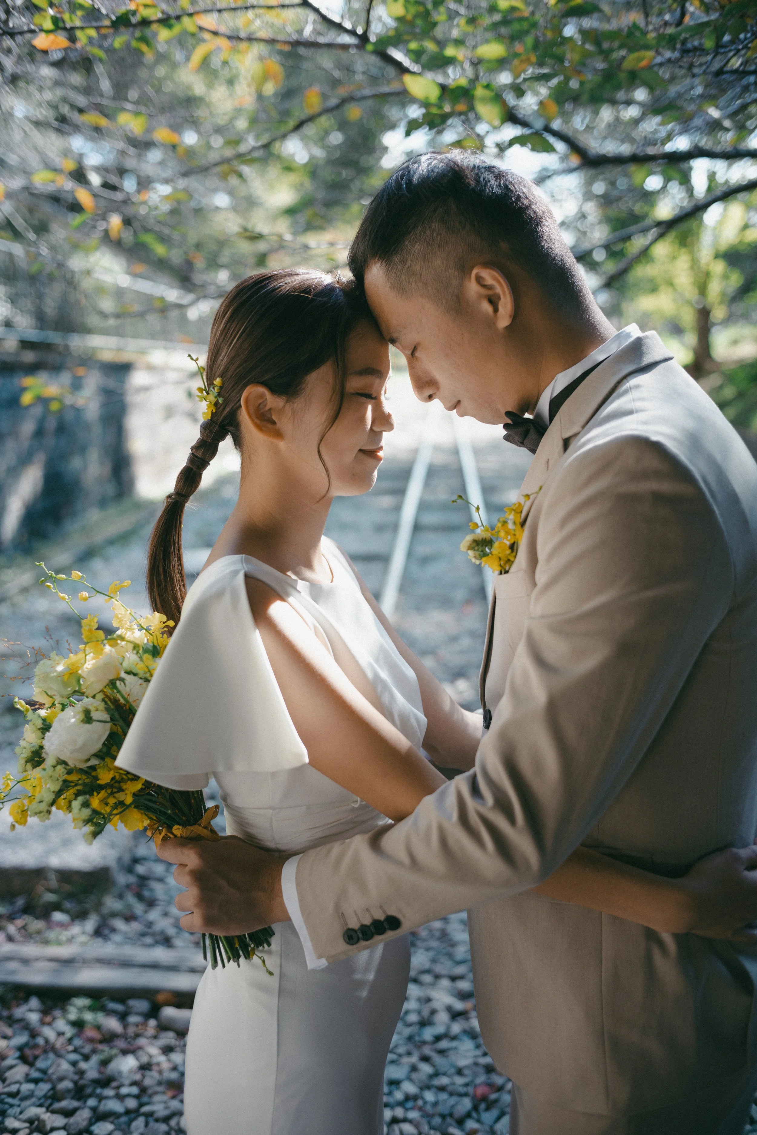 A bride and groom with their foreheads touching inside a wooded area, both holding a bouquet of yellow and white flowers, dressed in wedding attire.