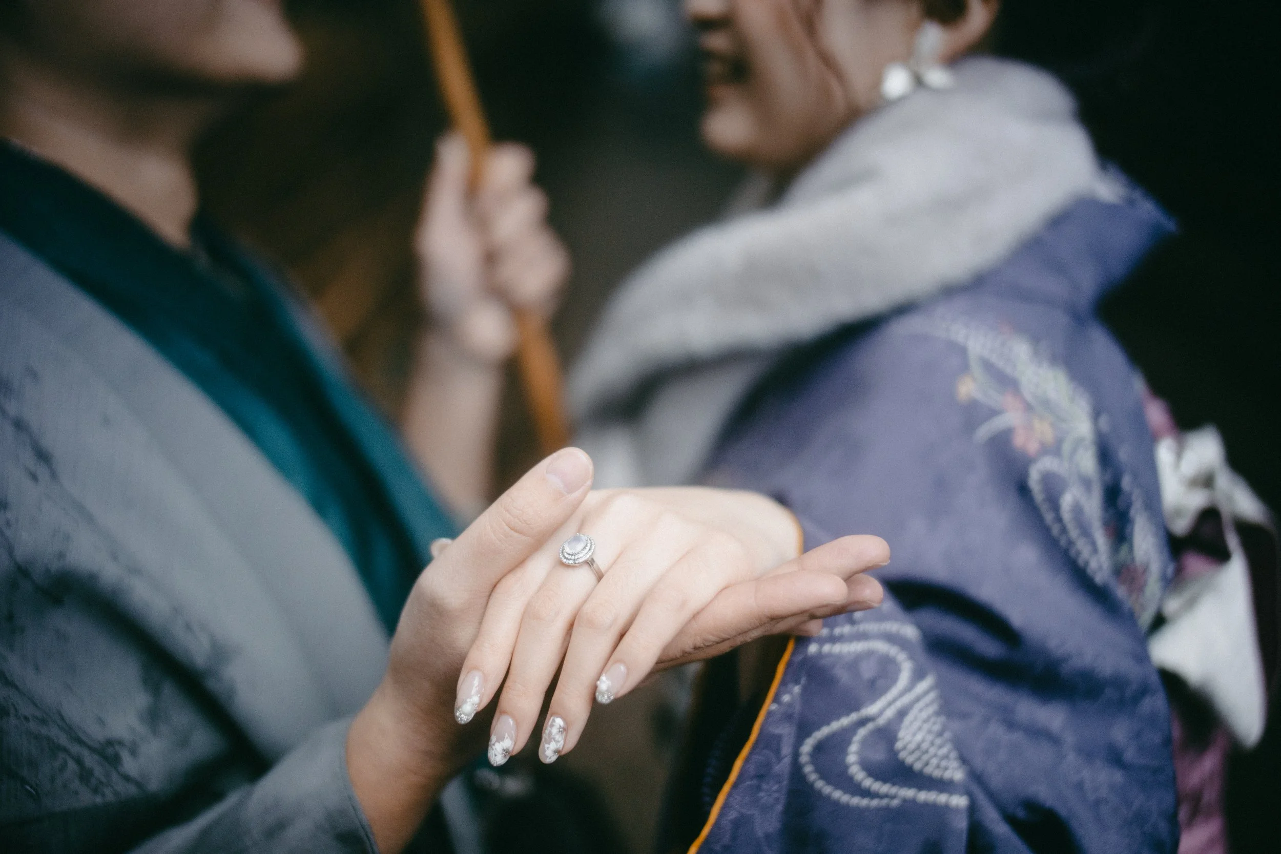 Close-up of two people holding hands, with a focus on a woman's hand wearing a diamond ring and decorated nails, while they face each other in a blurred background.