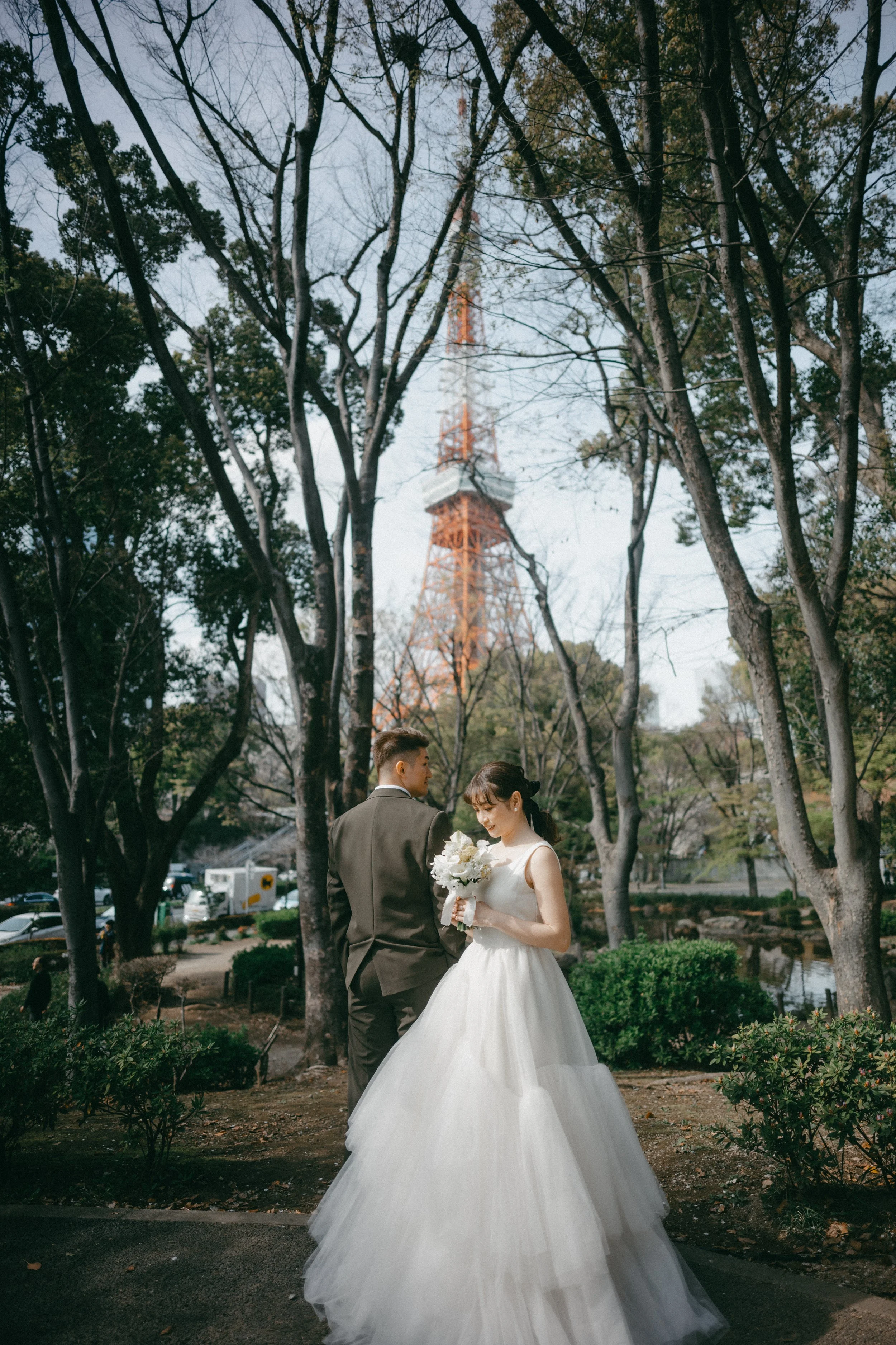 A bride in a white wedding gown holding a bouquet of flowers, standing next to a groom in a suit, in a park with trees and the Eiffel Tower in the background.