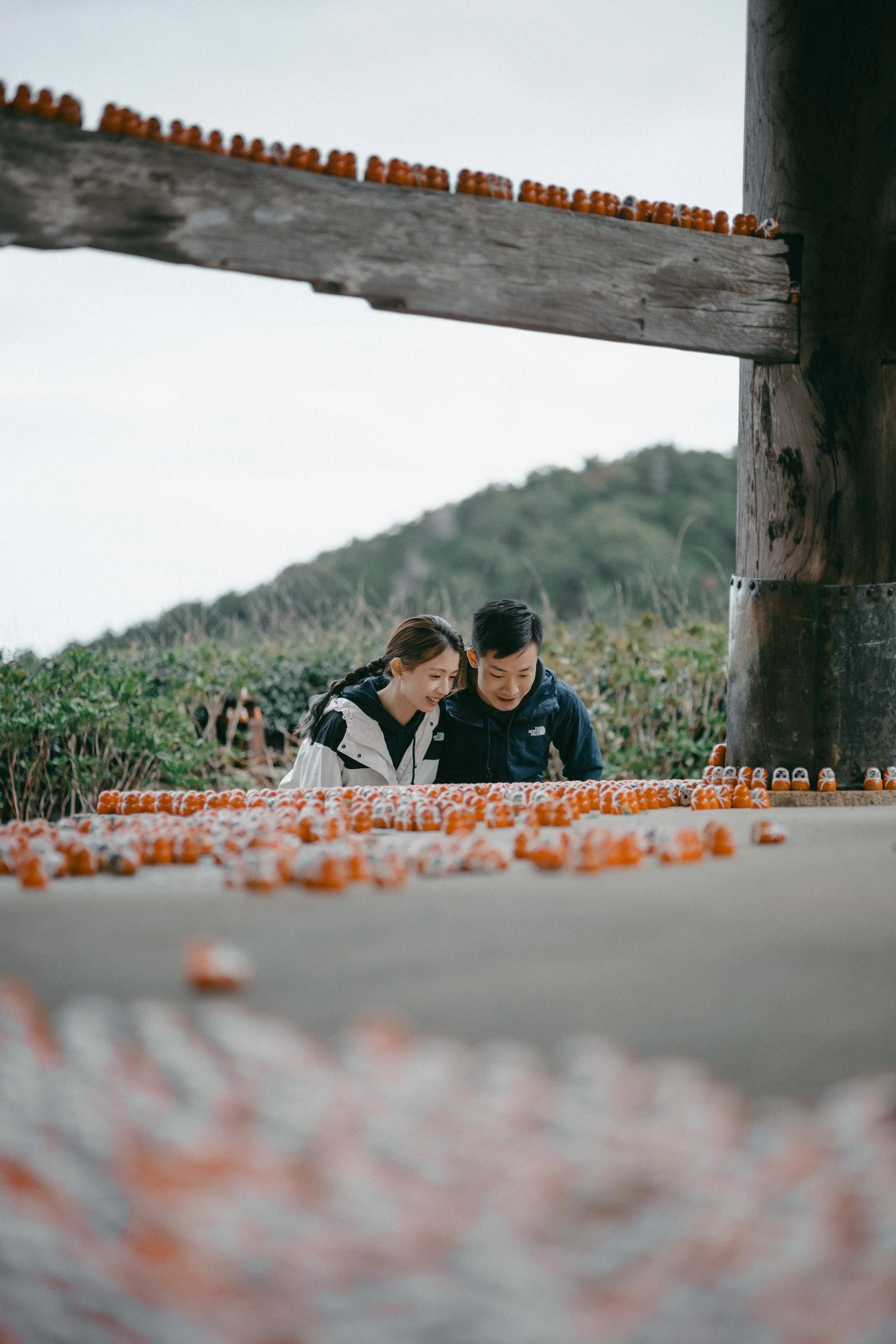 A young couple is smiling and looking at candy or small objects on the ground outdoors, with a large wooden structure and green hillside in the background.