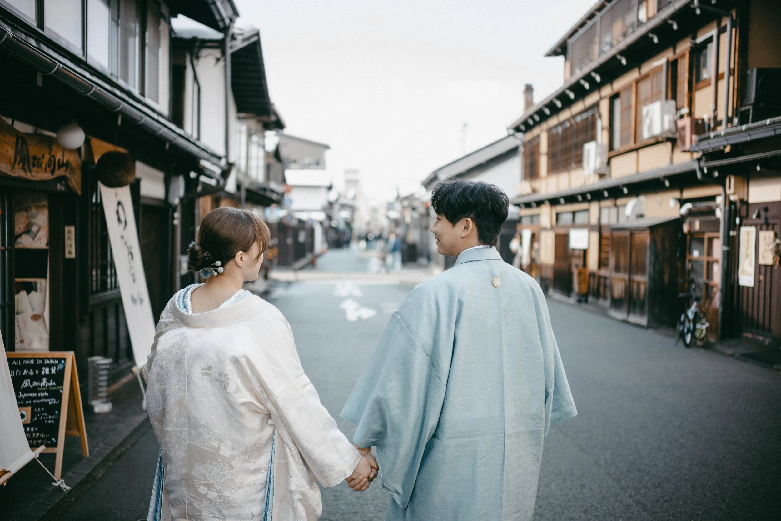 A couple dressed in traditional Japanese kimonos walking hand in hand down a street lined with Japanese shops and restaurants.