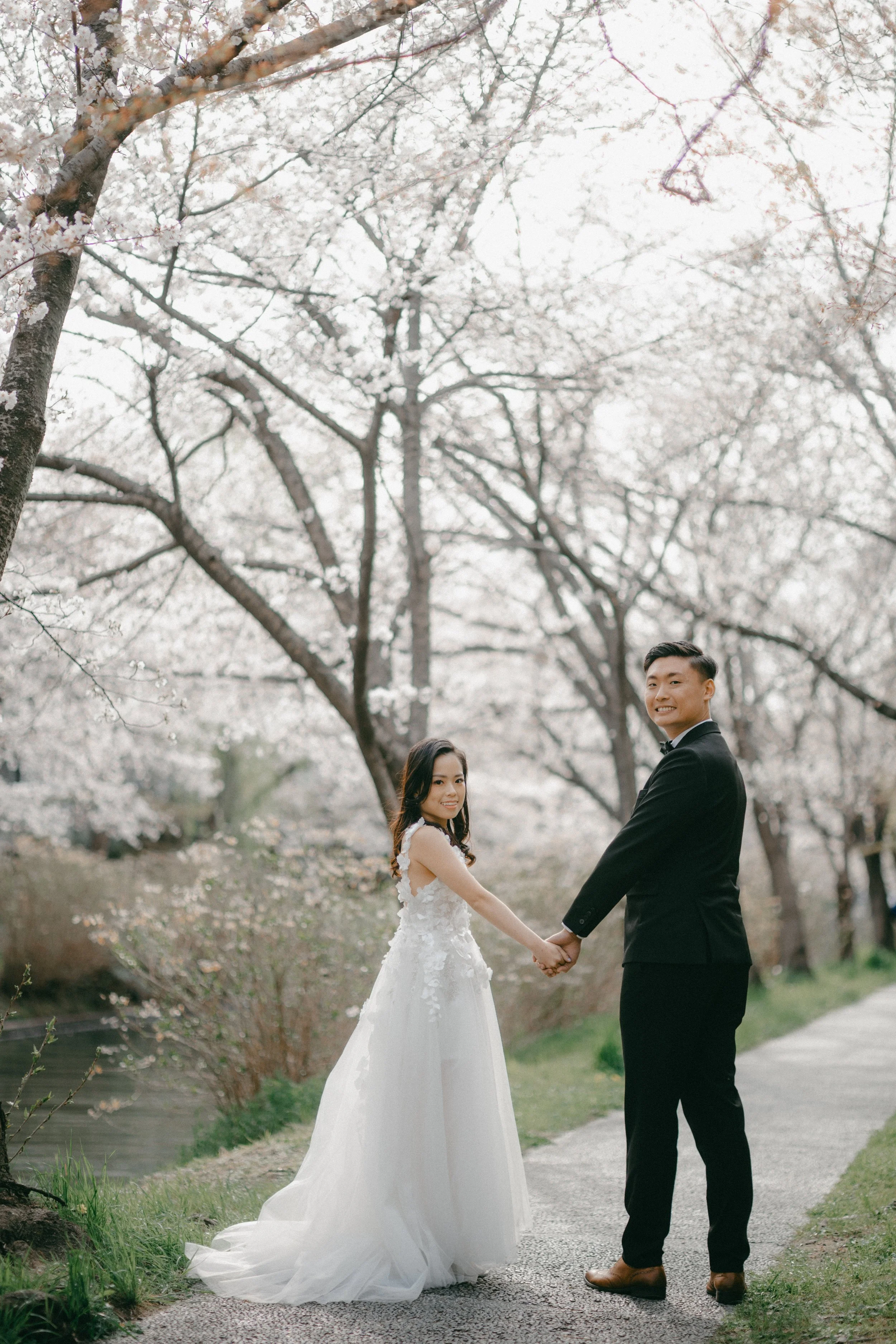 A bride and groom holding hands and smiling outdoors during spring, with blooming cherry blossom trees in the background.
