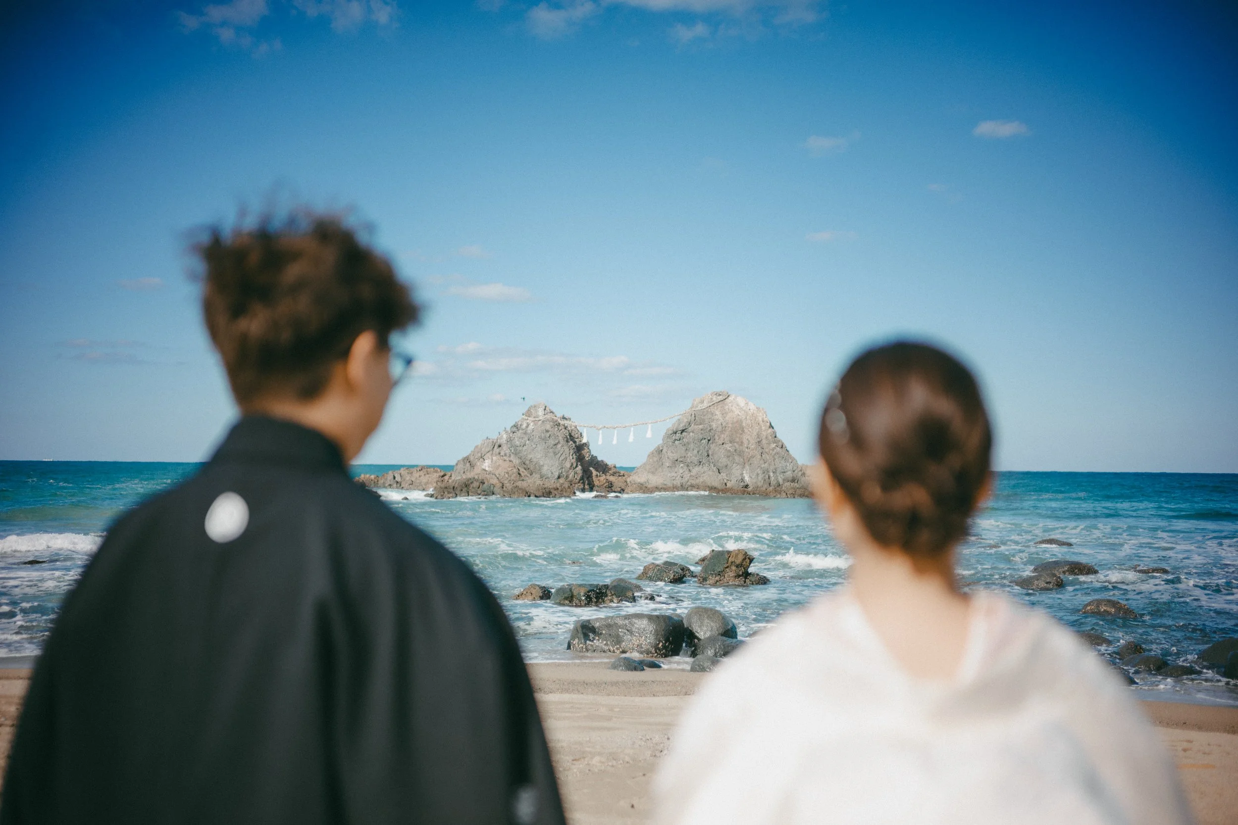 A man and woman sitting on a beach facing the ocean with two large rocks and a shimenawa in the background.