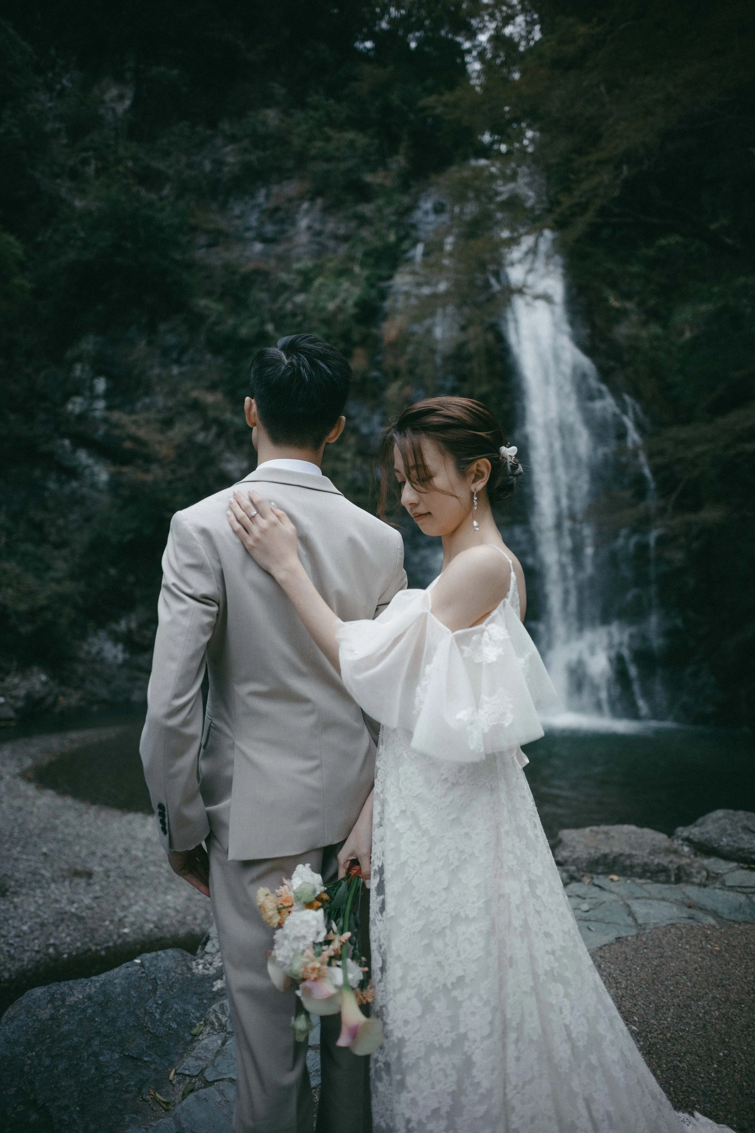 A bride and groom stand together by a waterfall in a lush, green outdoor setting, with the bride holding a bouquet of flowers.