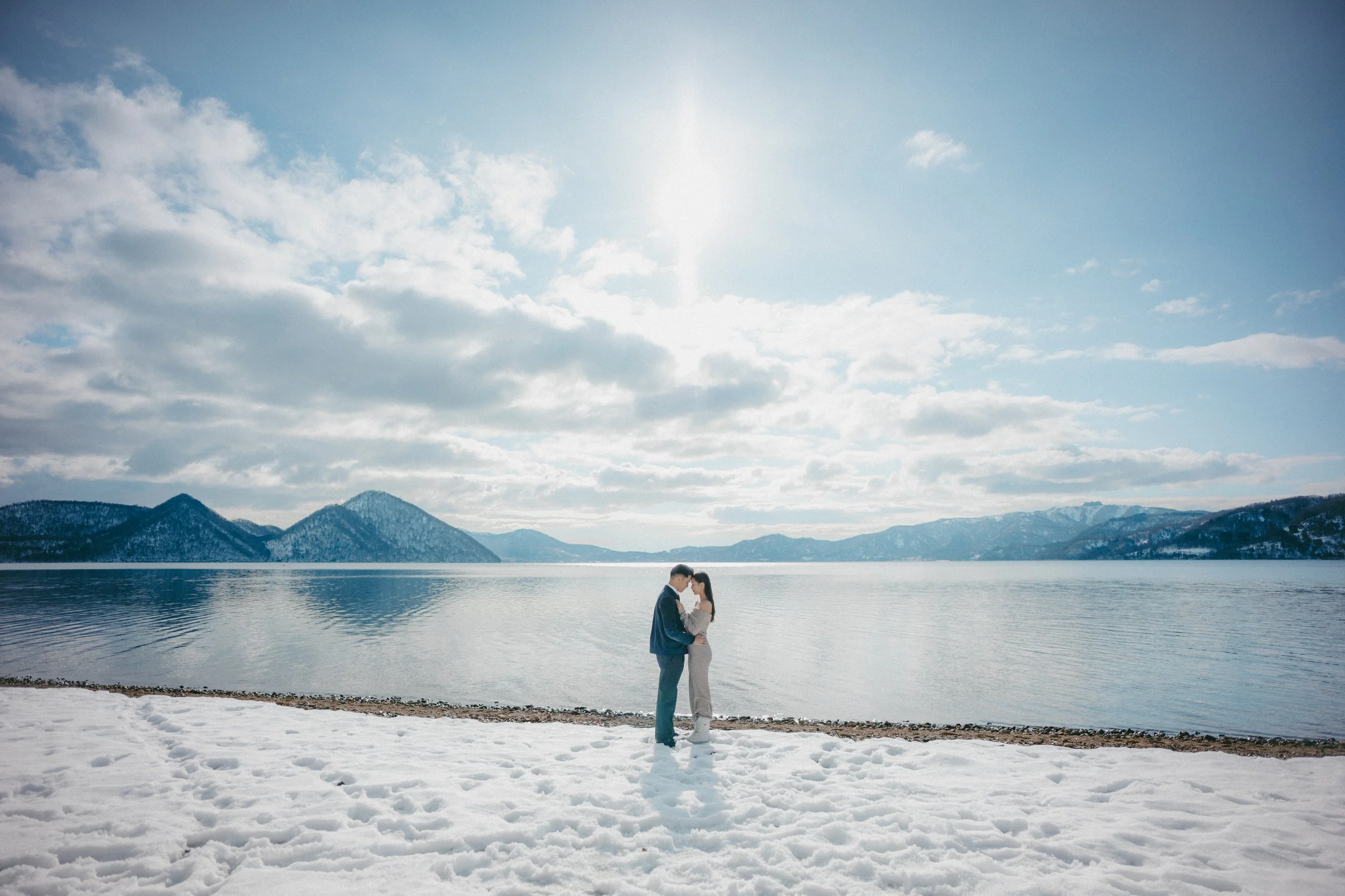 A couple stands close together on a snowy beach, with mountains and a lake in the background under a partly cloudy sky.