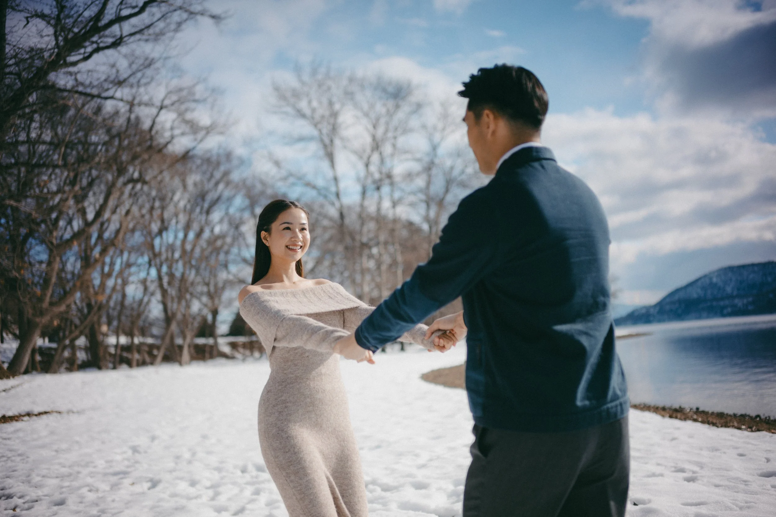 A couple is holding hands and dancing on snow-covered ground near a lake with mountains in the background during daytime. The woman is smiling and wearing a beige dress, while the man wears a dark jacket.