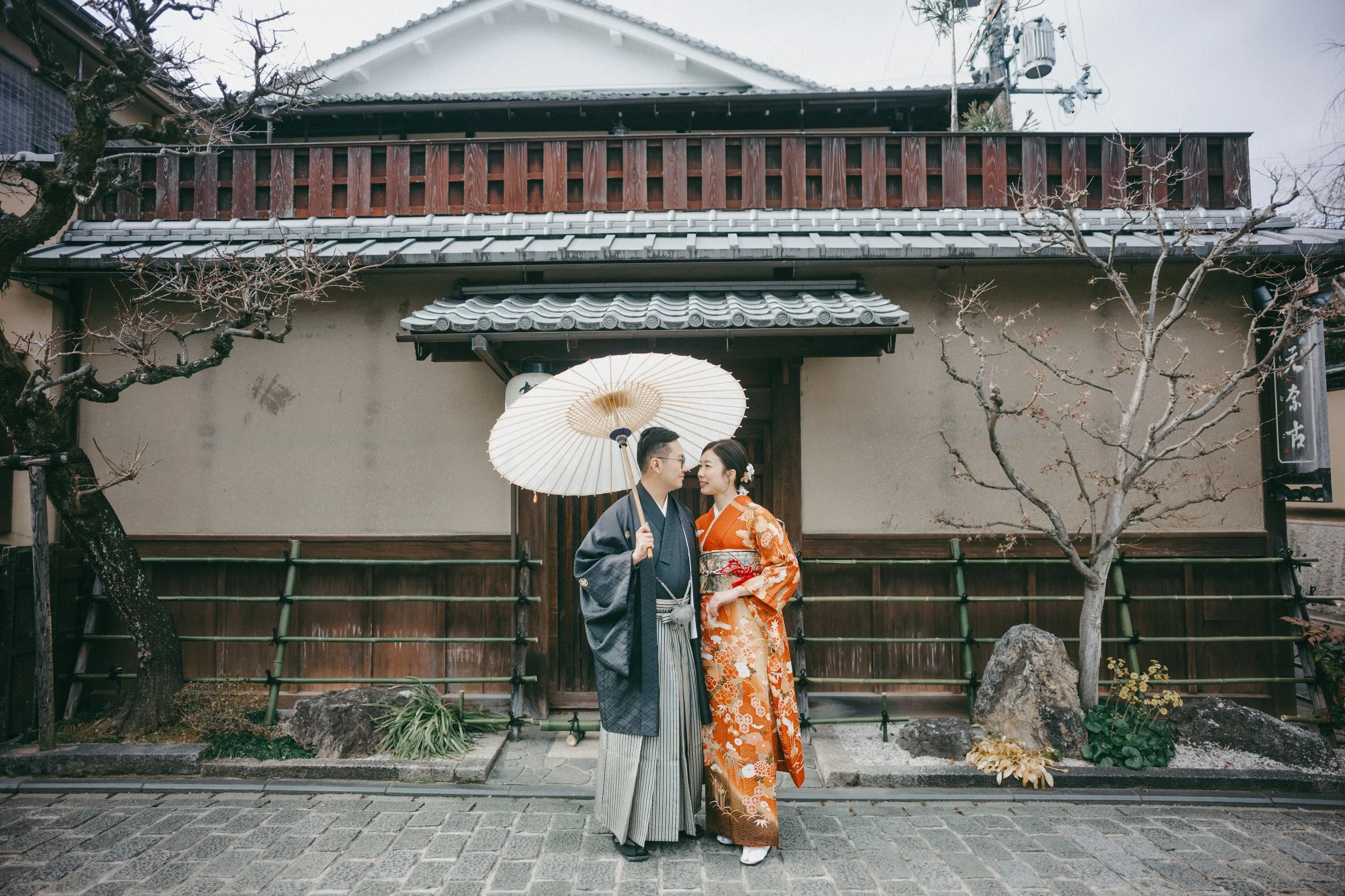 Couple dressed in traditional Japanese kimonos standing in front of a Japanese building, with the man holding a parasol over the woman.