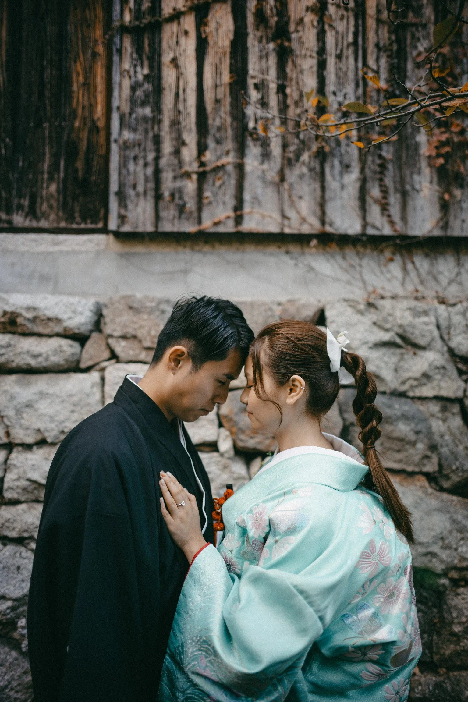 A couple dressed in traditional Japanese wedding attire, standing close with foreheads touching in front of a stone wall.