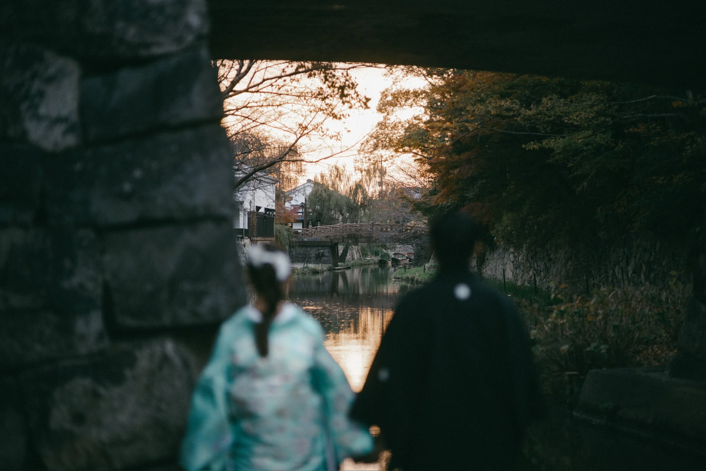 Two people, a woman in a kimono and a man in a dark coat, sitting on rocks under a bridge, overlooking a river with trees and houses during sunset.