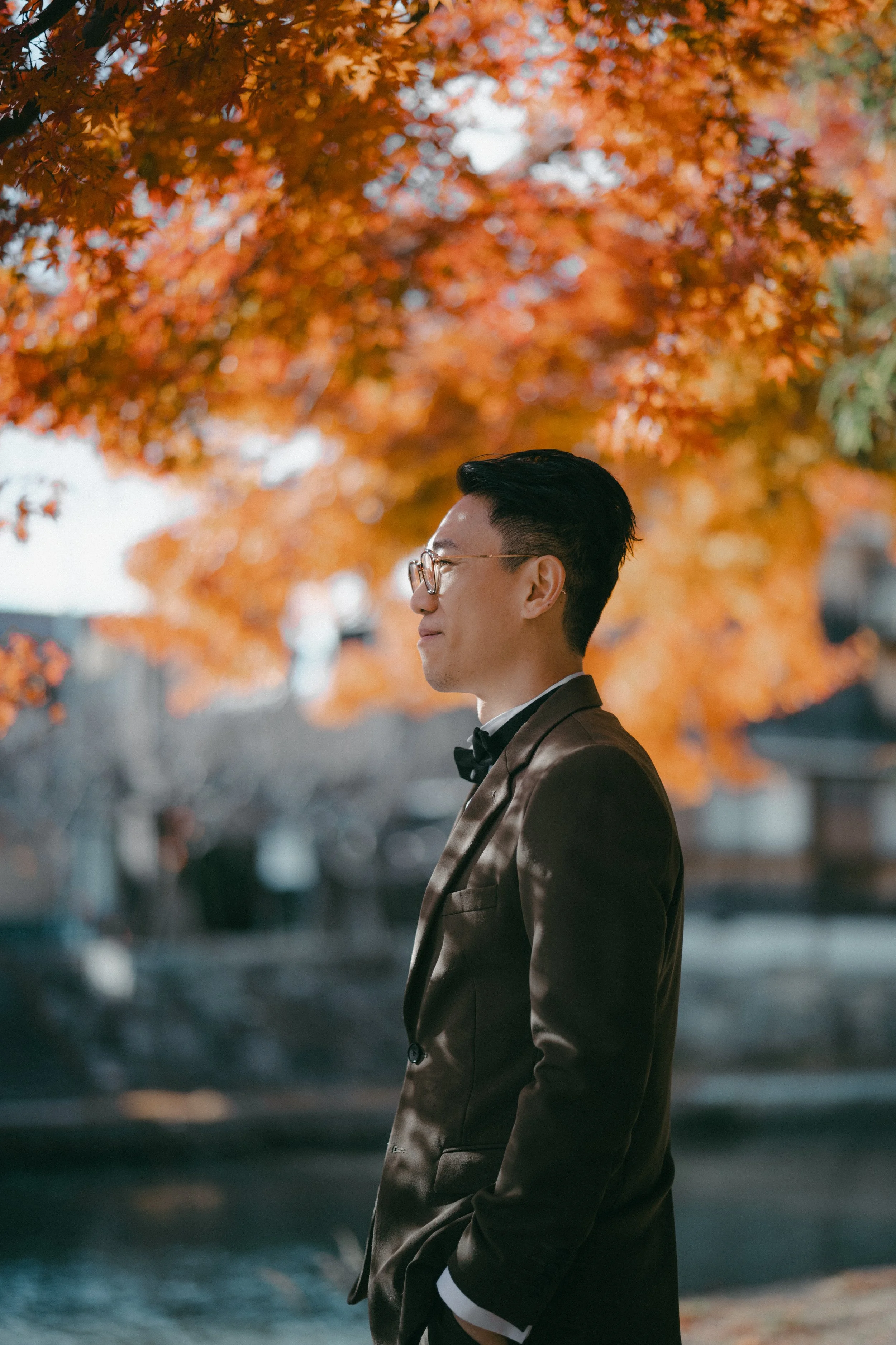 A man in a tuxedo with glasses standing outdoors during autumn, with orange and red leaves on trees in the background.