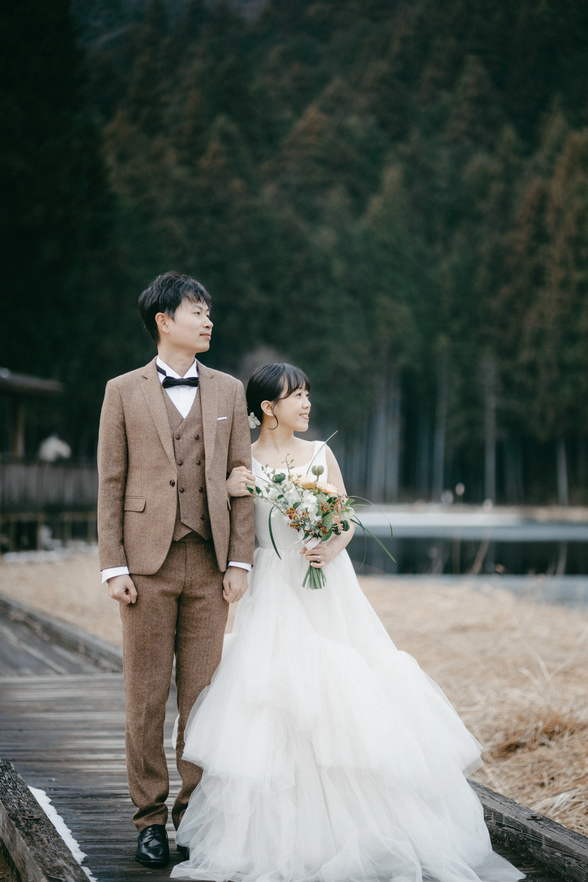 A bride and groom standing together on a wooden pathway outdoors, with the bride holding a bouquet of flowers and the groom in a brown suit, against a forested background.