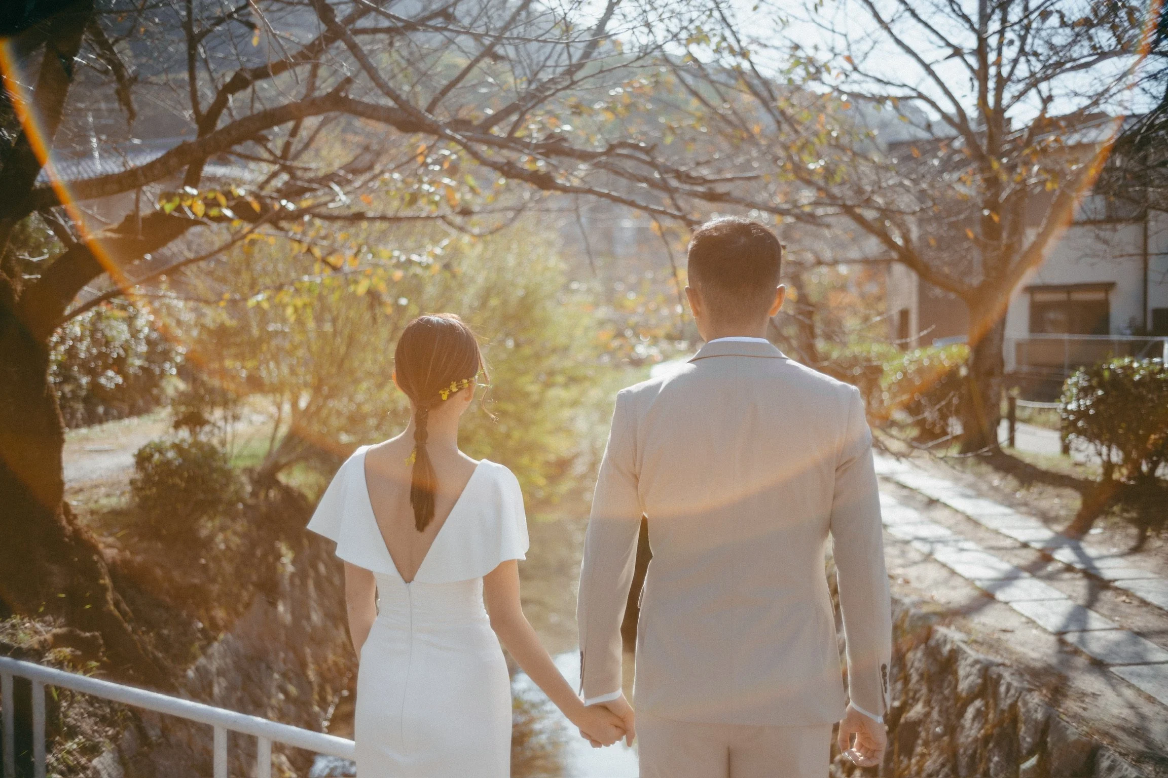A bride and groom holding hands and walking along a small river or stream on a sunny day with trees and buildings in the background.