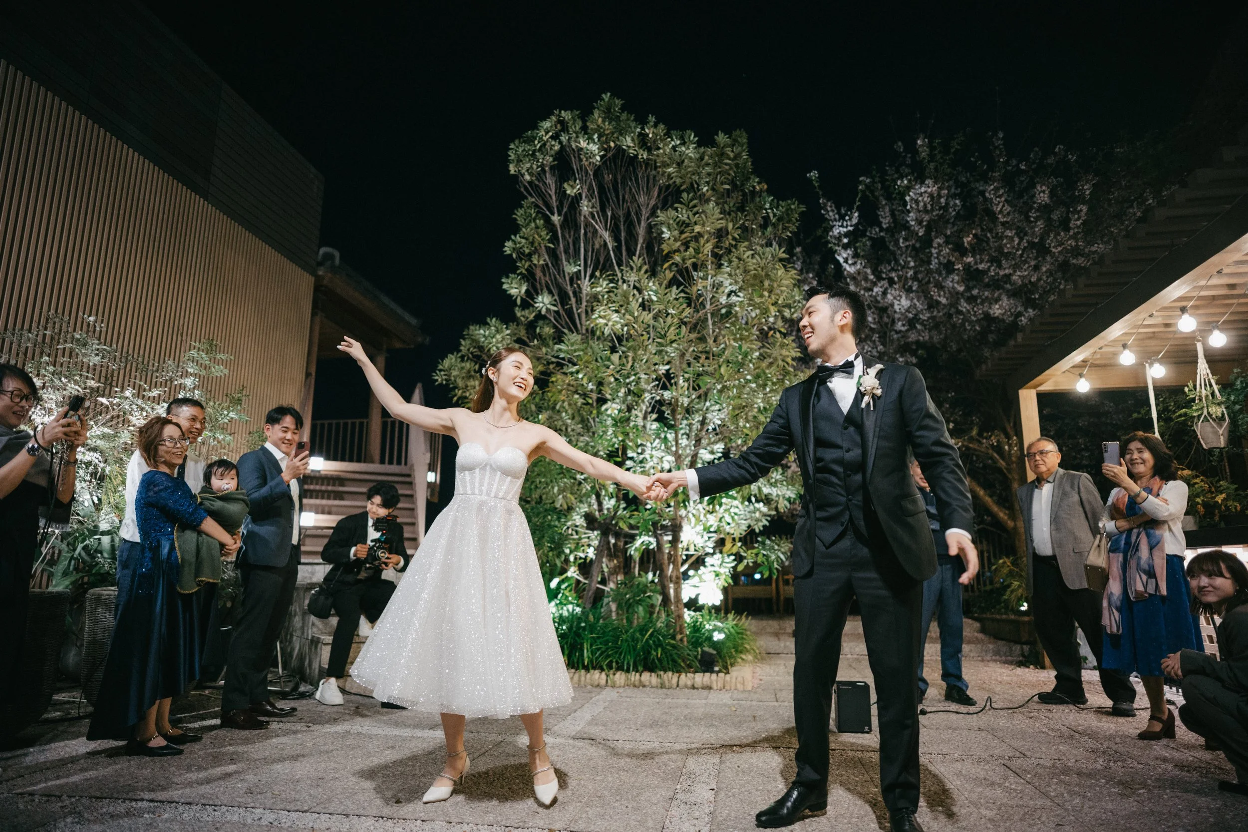 A bride and groom dancing happily at their wedding reception outdoors at night, surrounded by friends and family taking photos and videos.