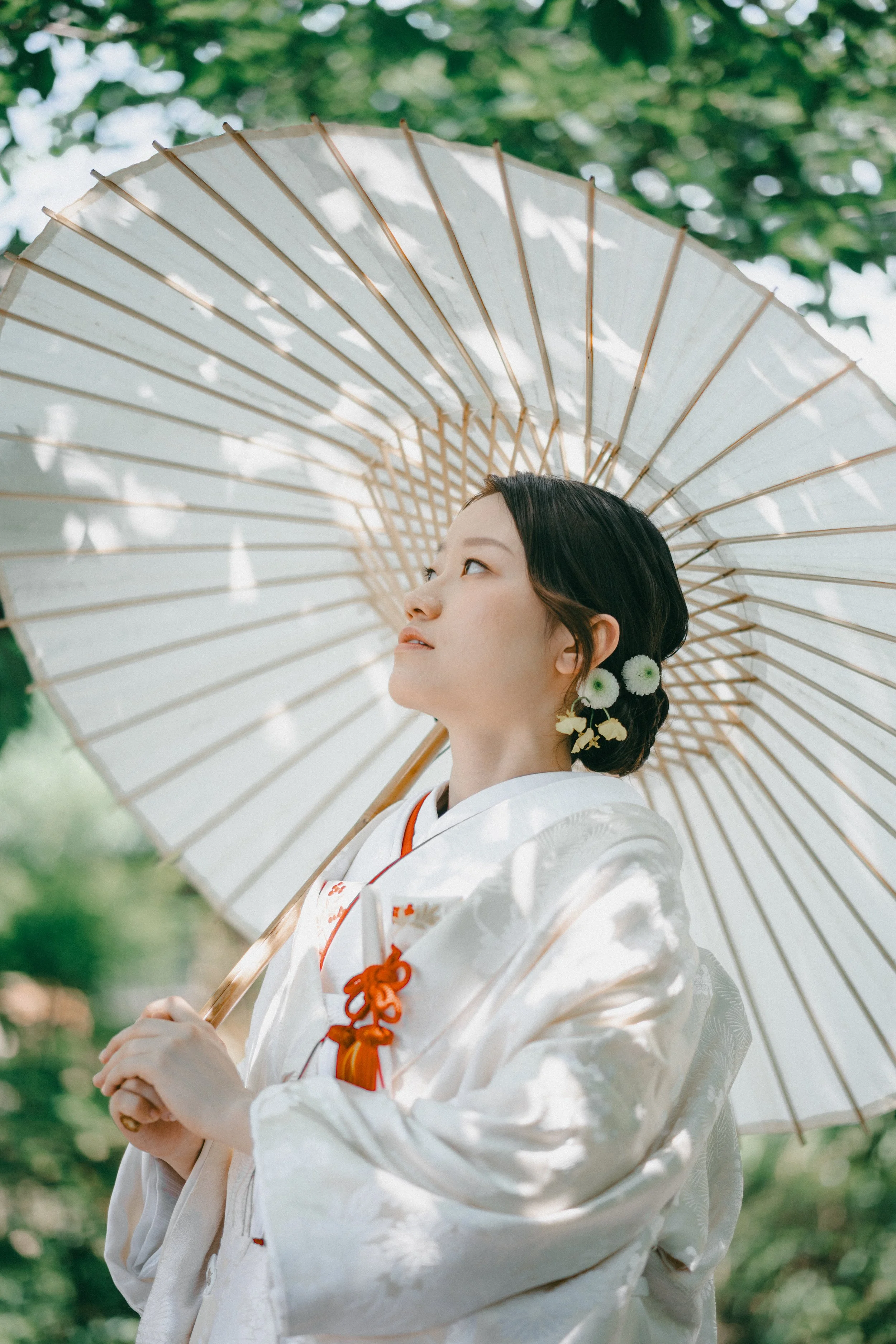 A young woman dressed in traditional Japanese attire, holding a white parasol, standing outdoors with green foliage and sunlight filtering through the trees.