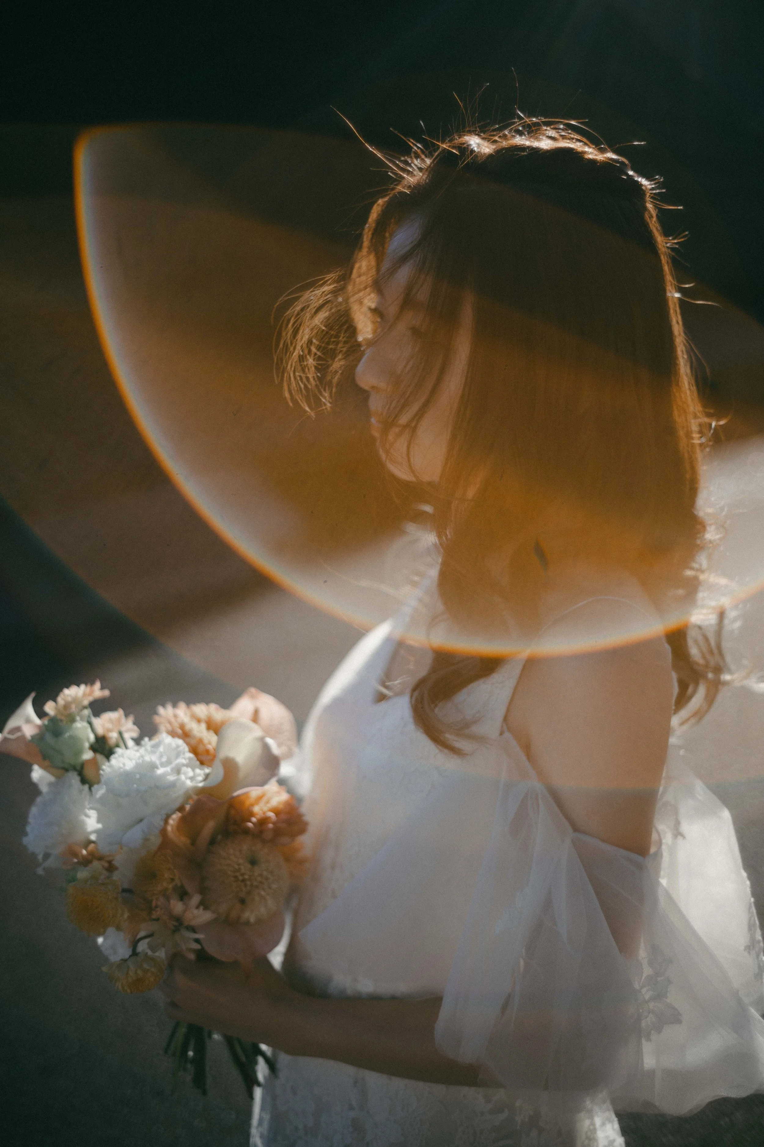 A woman in a white dress holding a bouquet of flowers, wearing a large sunhat, with sunlight creating a rim light around her profile.