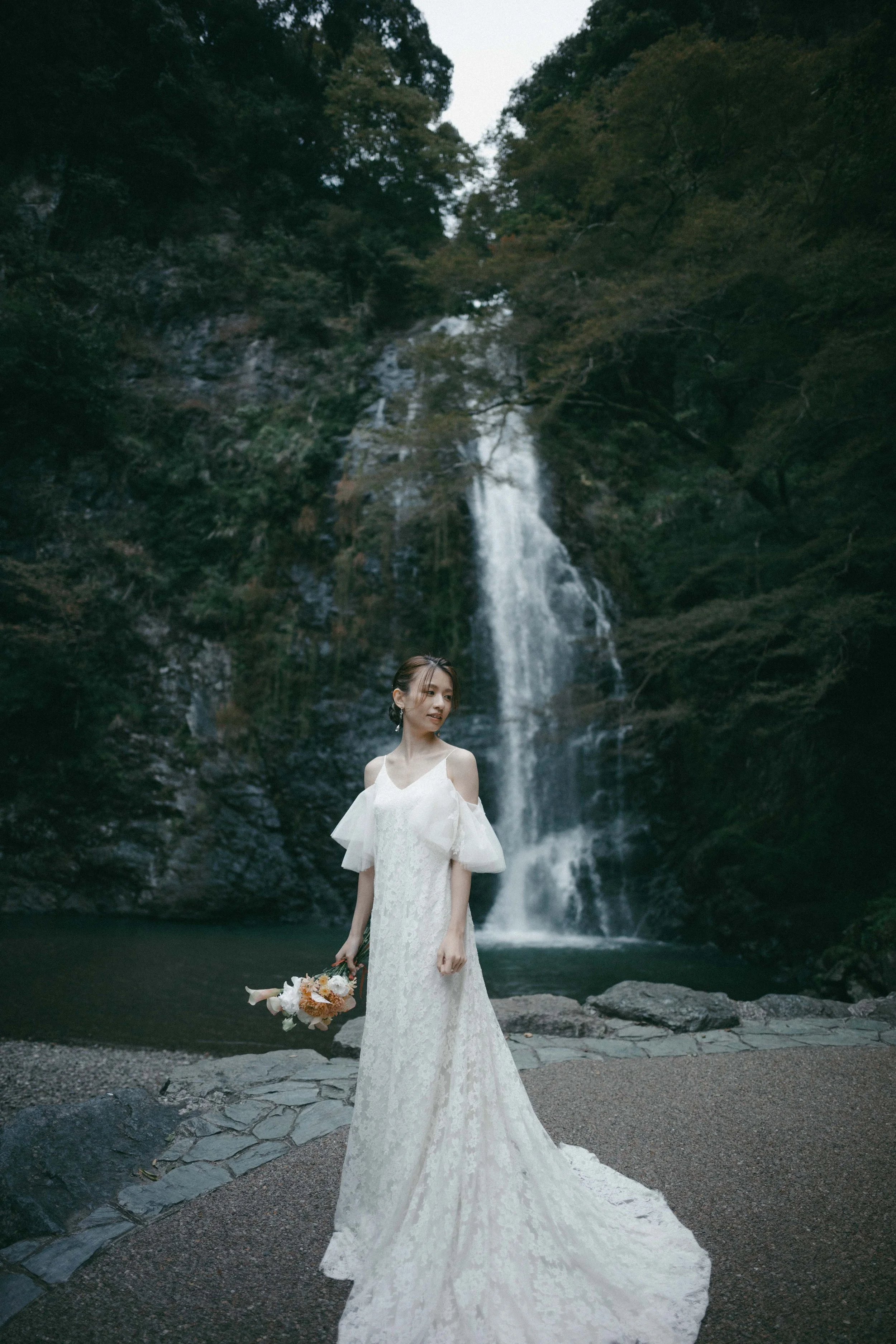 A woman in a white wedding dress holding a bouquet stands in front of a waterfall surrounded by trees.