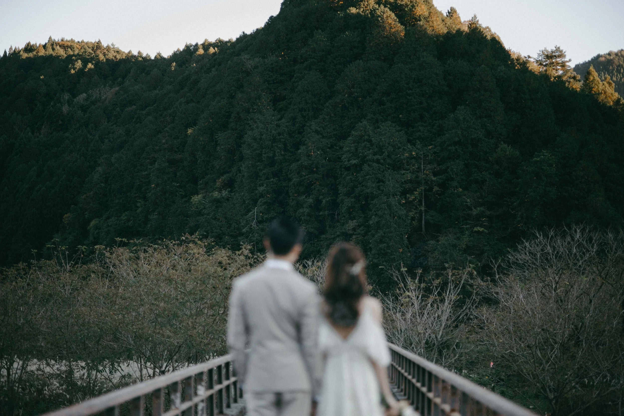 A couple dressed in wedding attire, walking hand in hand on a bridge towards a mountain covered with dense forest, during sunset.