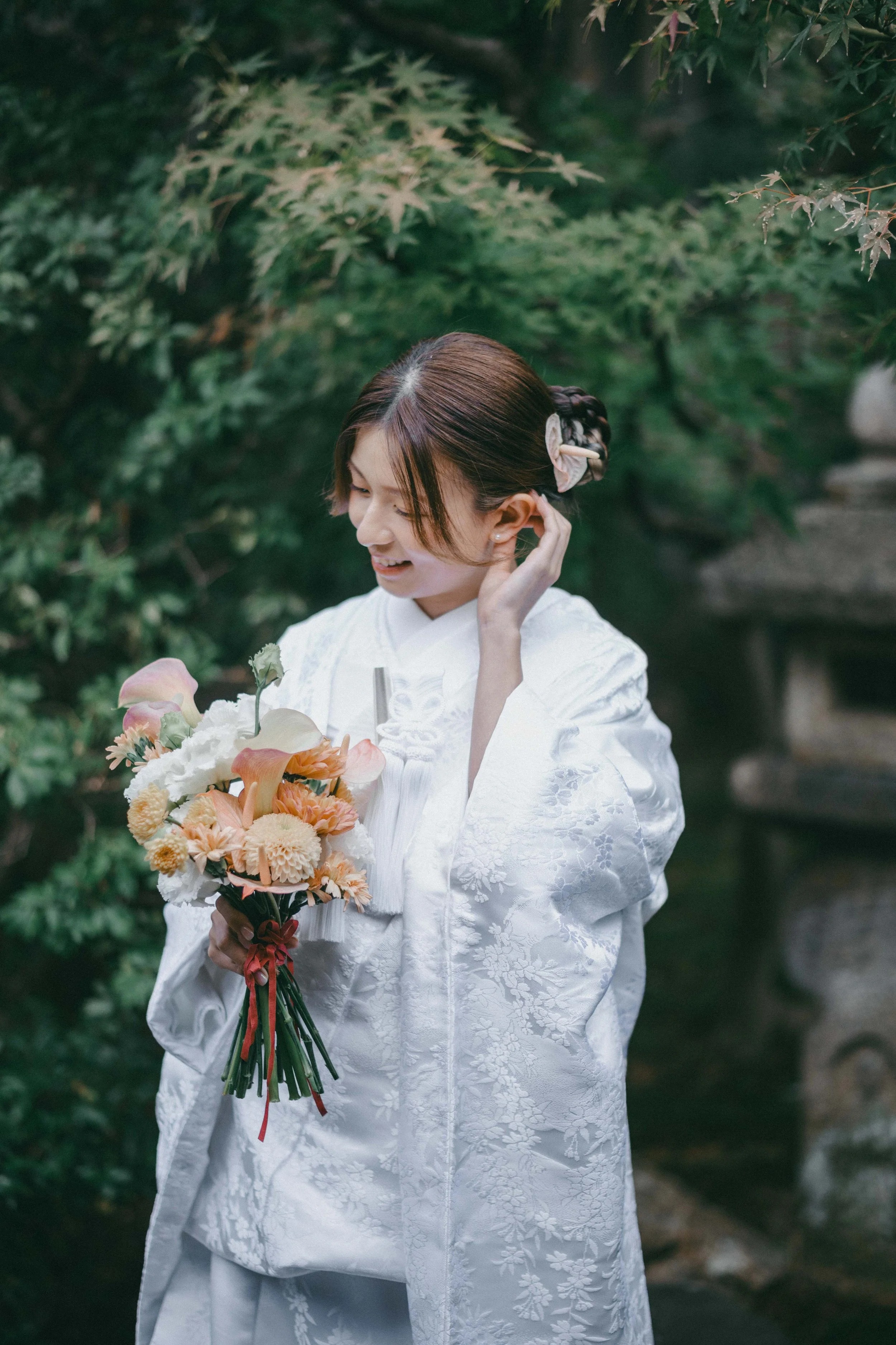 A smiling woman dressed in a white traditional kimono holding a colorful bouquet of flowers outdoors among green foliage.