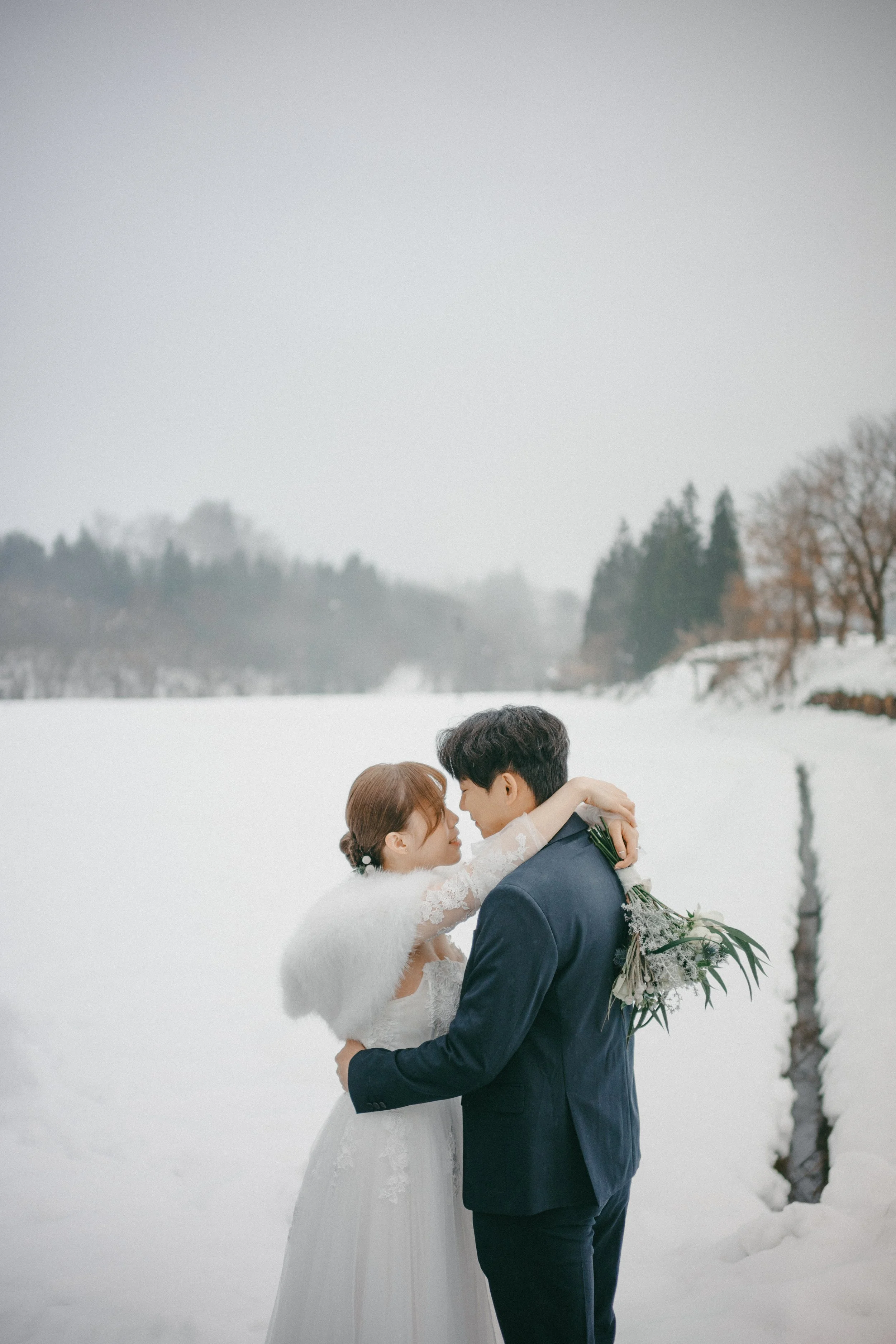 A bride and groom in wedding attire embrace in a snowy landscape, with snow-covered ground and distant trees in the background.