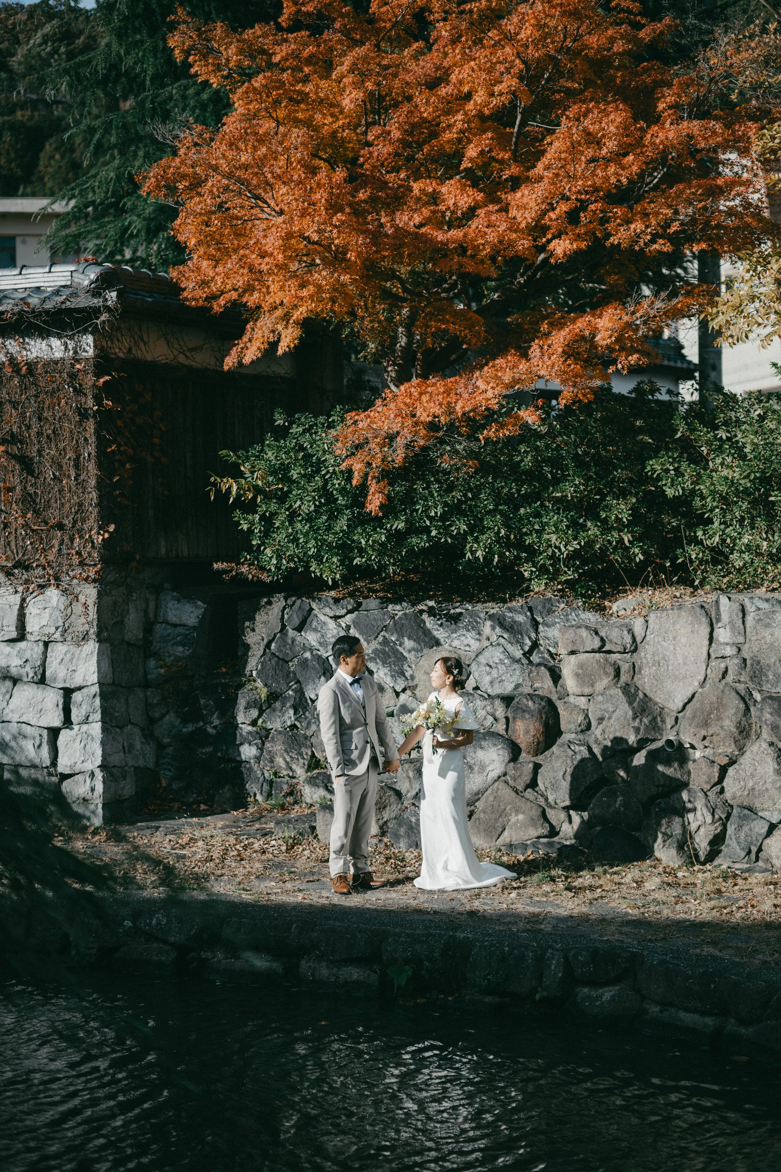 A bride and groom holding hands outdoors with autumn foliage and a stone wall in the background.