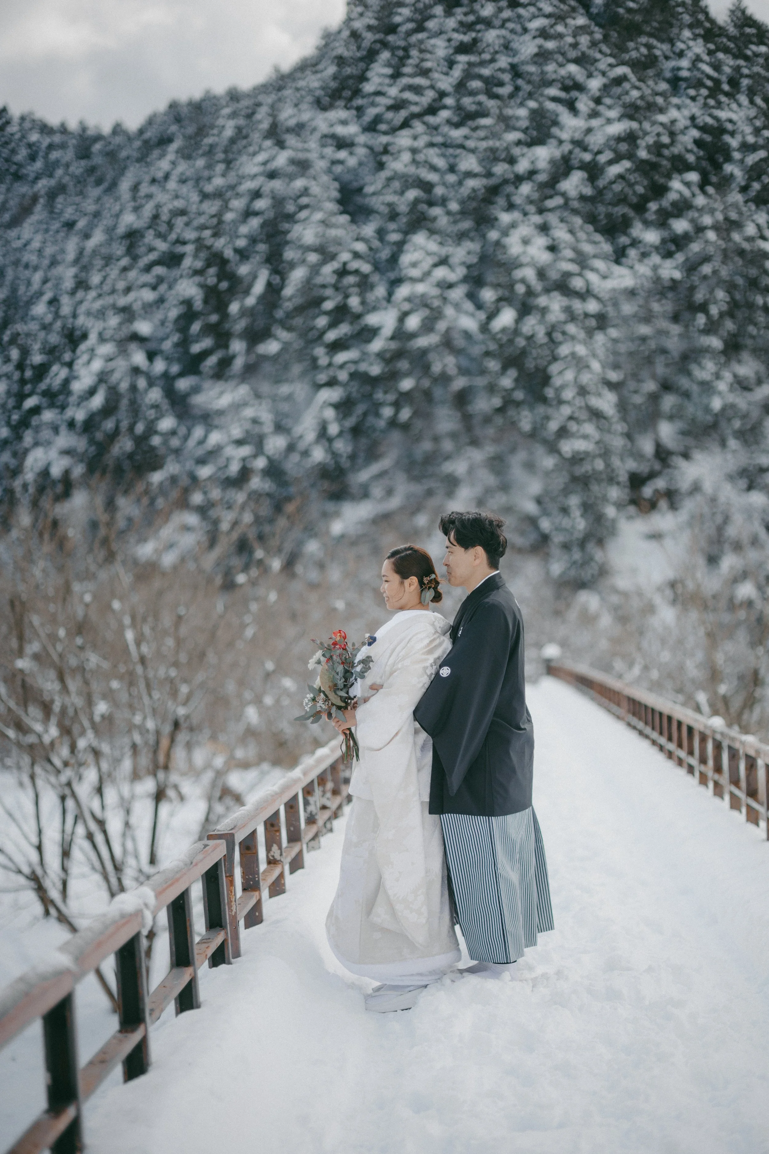 A couple in traditional Japanese wedding attire standing on a snow-covered bridge with snowy trees and mountains in the background.