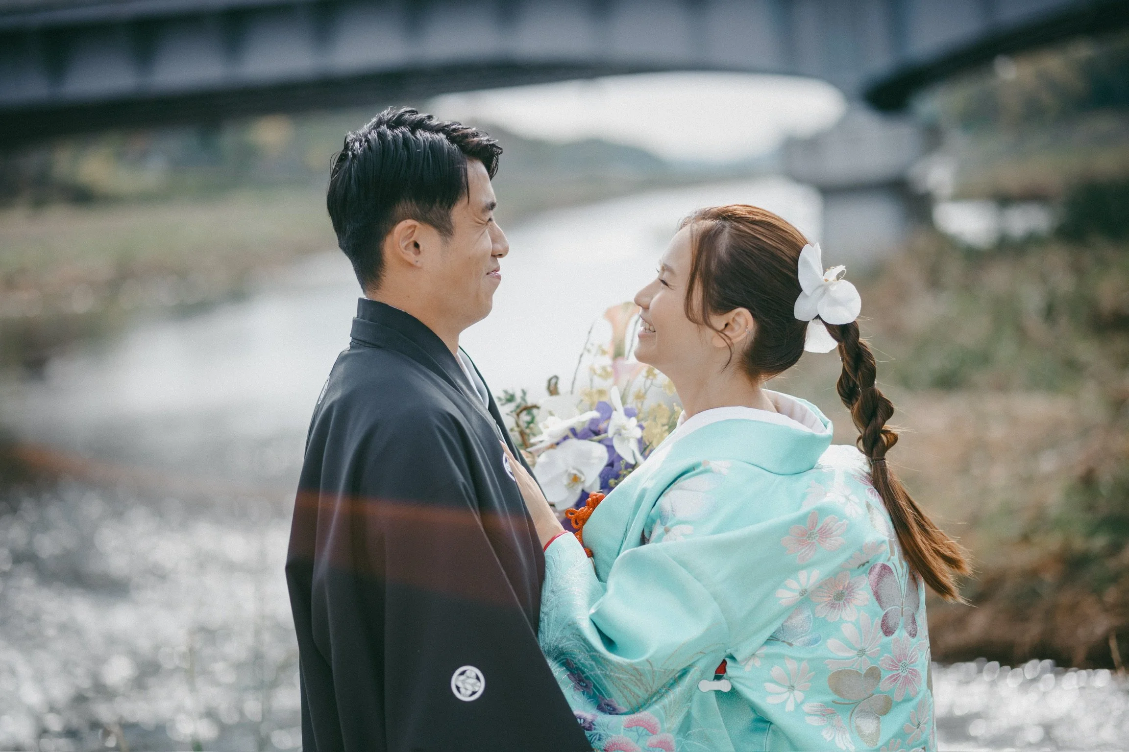 A couple dressed in traditional Japanese clothing, smiling and looking at each other near a river with a bridge in the background.