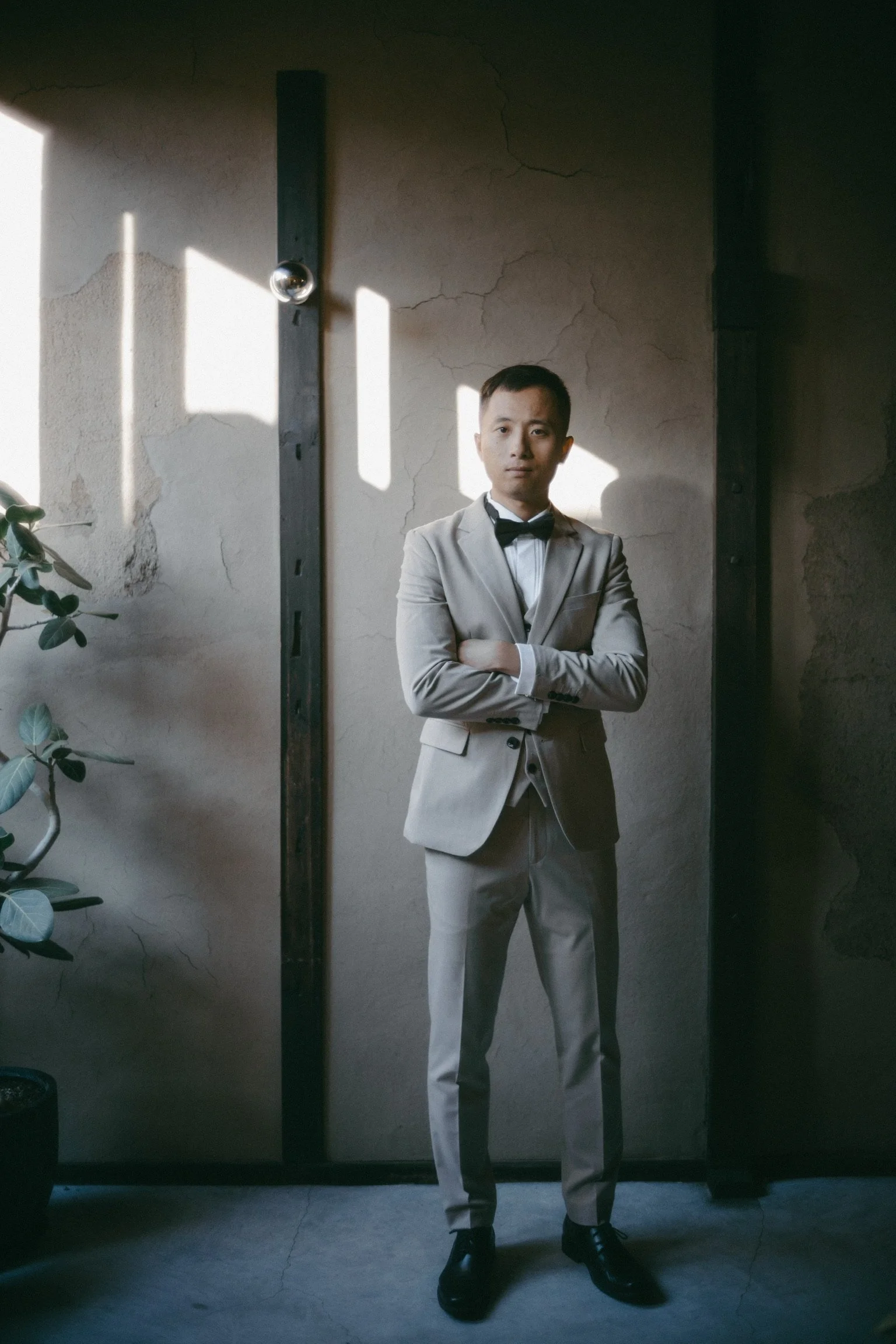A man in a gray suit with a bowtie standing with crossed arms in front of a textured wall with sunlight and shadow patterns.
