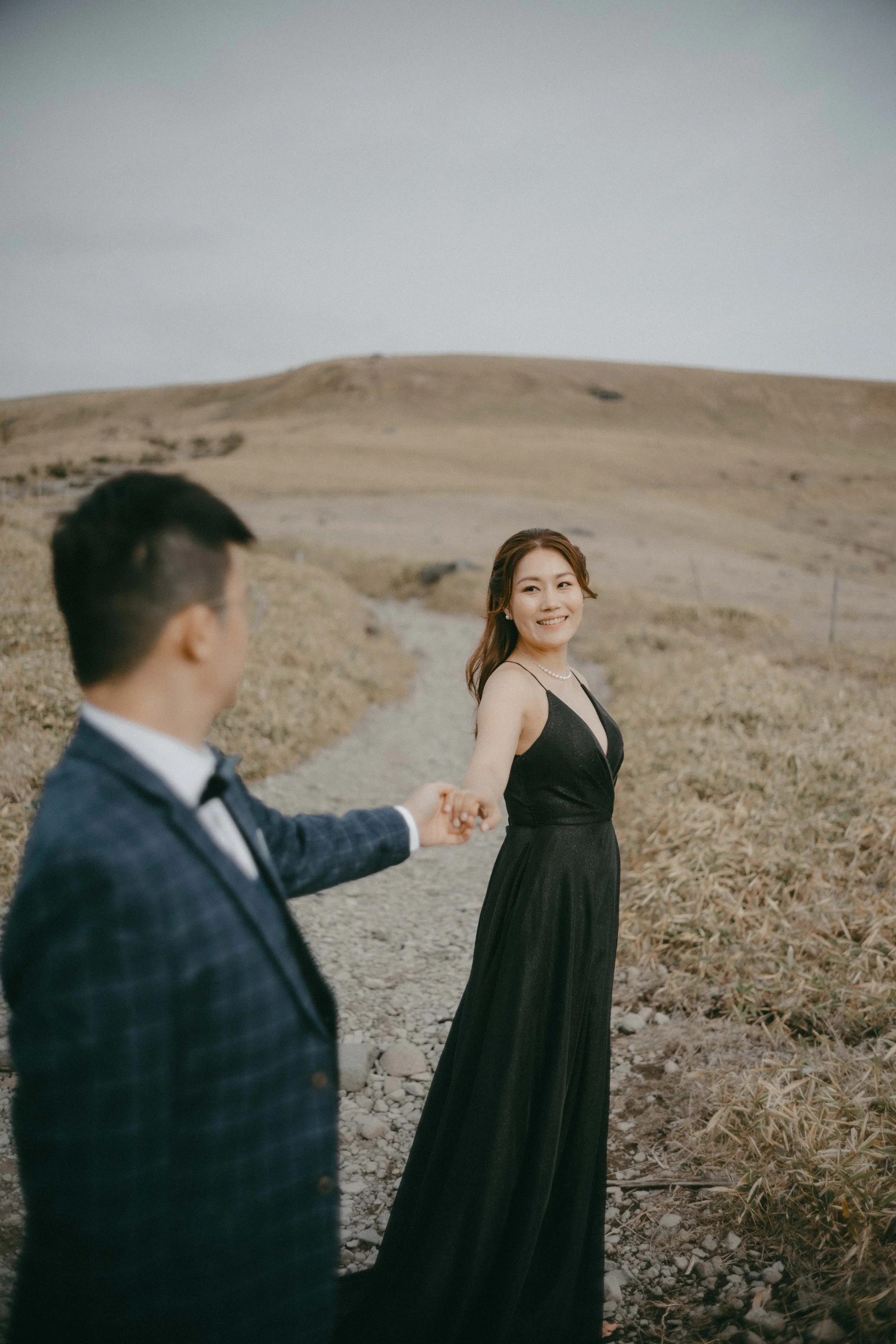A woman in a black dress holding hands with a man in a suit on a dirt path in a barren, hilly landscape.