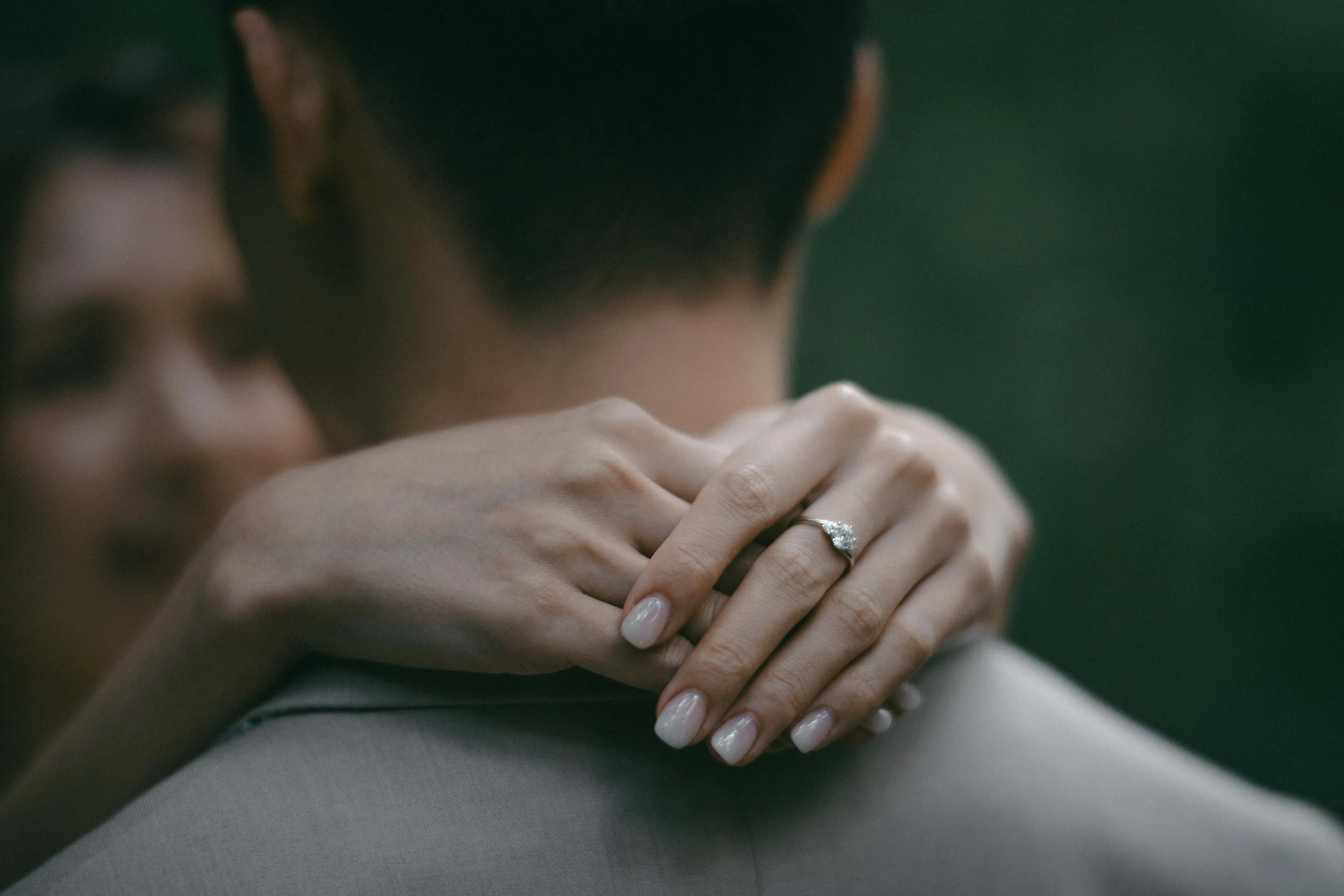 A woman resting her head on a person's shoulder, showing her engagement ring, with a blurred background.