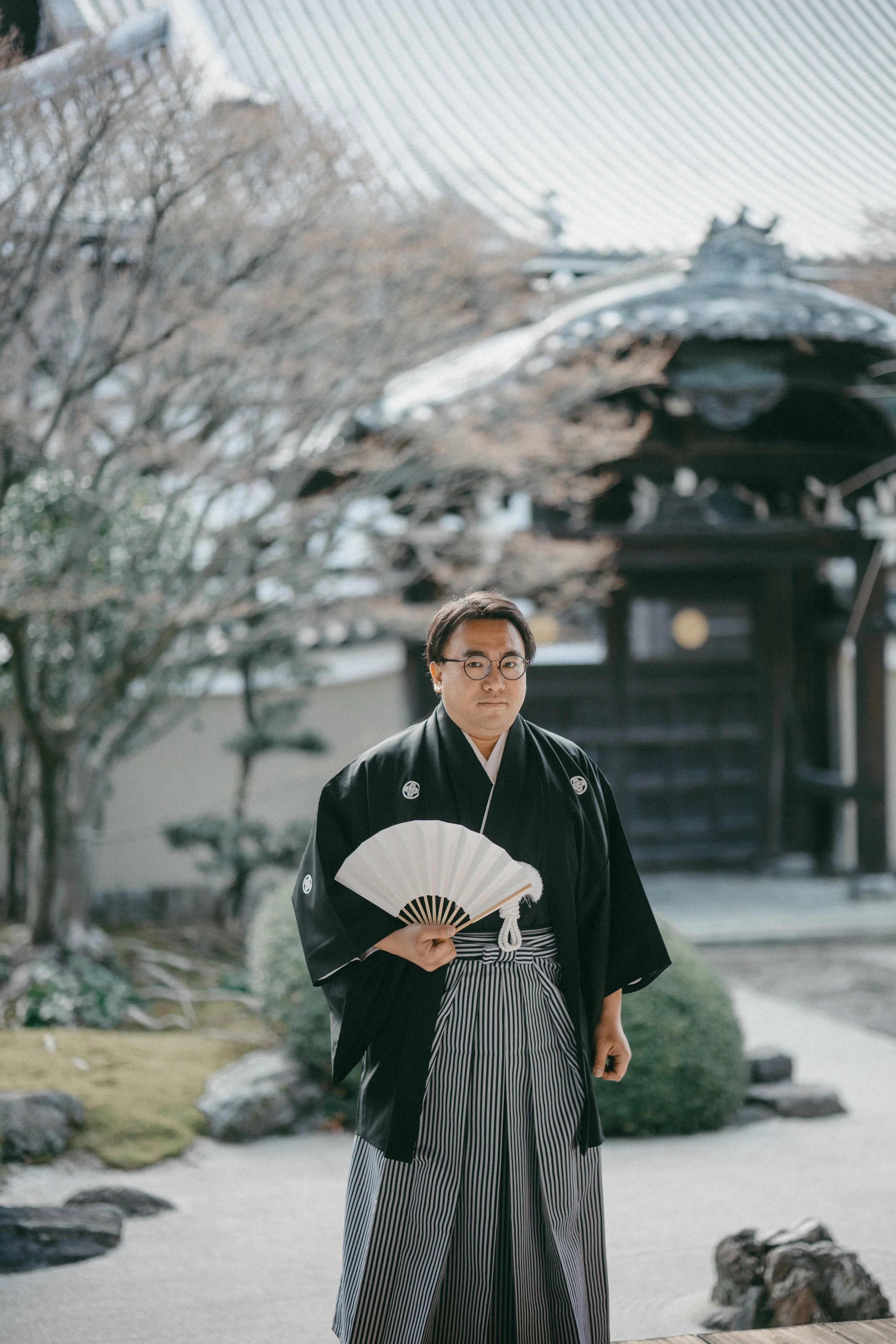 A man in traditional Japanese attire holding a fan, standing in a Japanese garden.