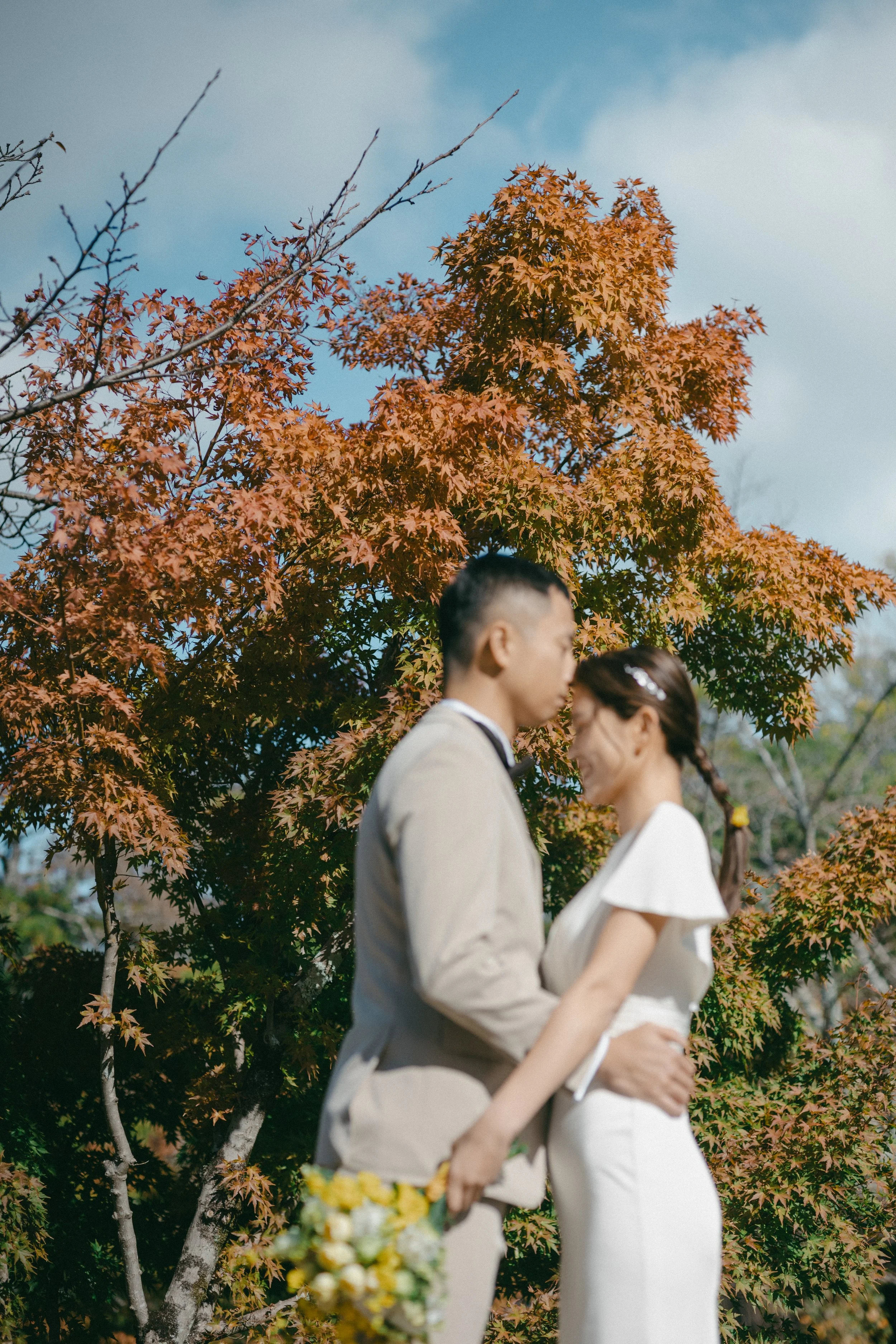 A couple dressed in wedding attire embracing outdoors during autumn, with orange and green leaves on trees in the background.