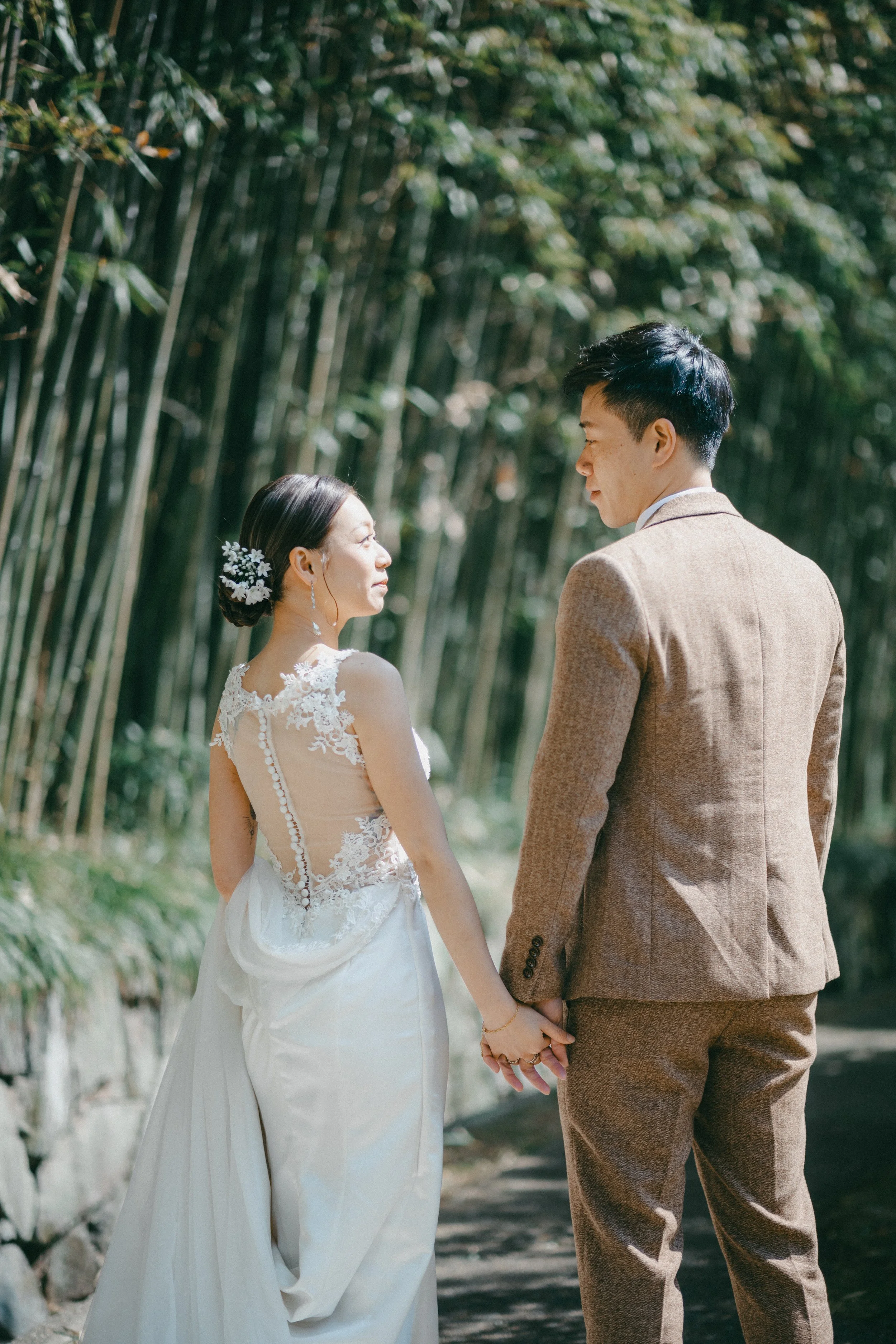 A bride and groom holding hands and looking at each other outdoors among bamboo trees, with sunlight filtering through.