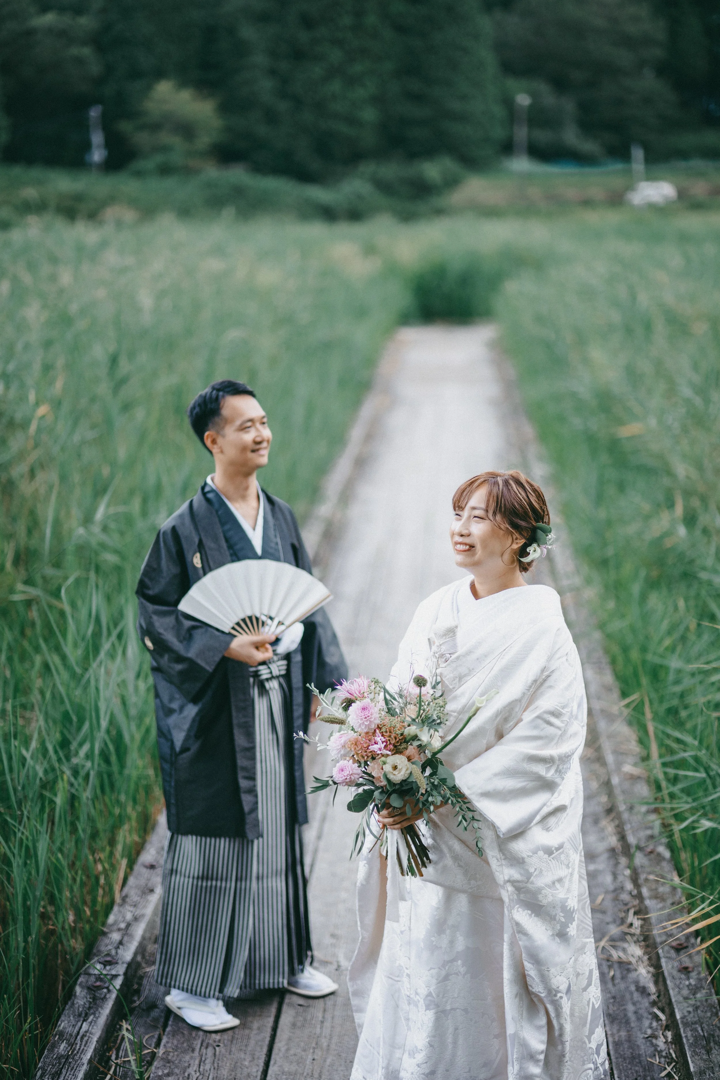 A bride and groom in traditional Japanese wedding attire standing on a wooden pathway surrounded by green fields, smiling and holding flowers.