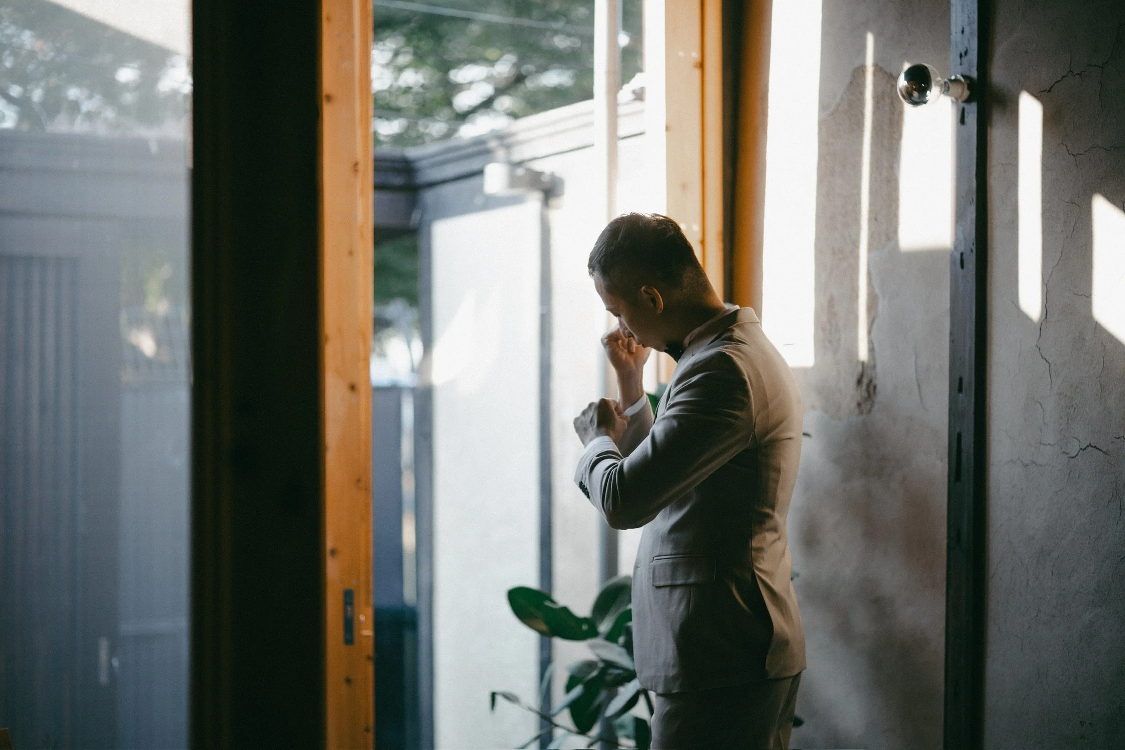 A man in a light-colored suit adjusting his cufflink inside a room with natural light coming through a window, with a potted plant nearby.