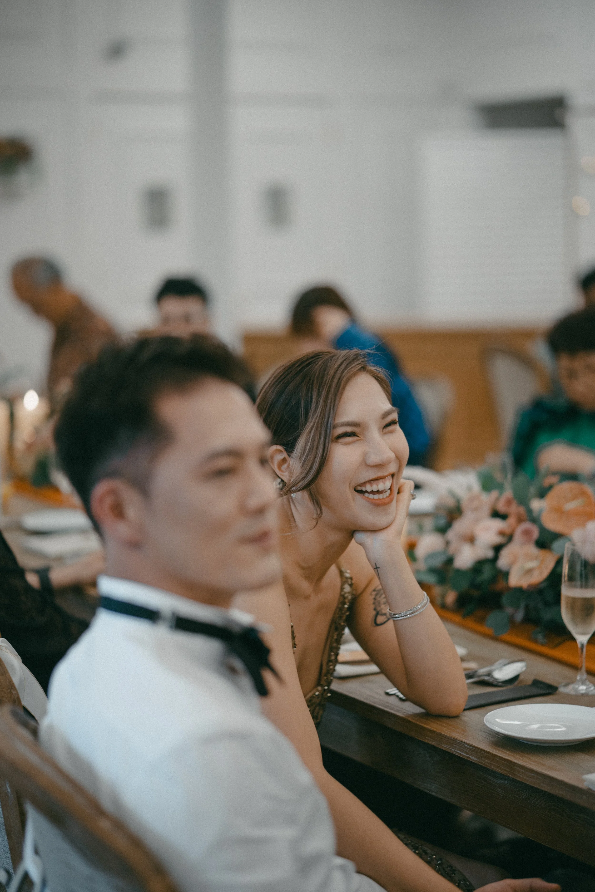 A woman laughing at a formal event, seated at a table with a man, surrounded by other people, floral centerpiece, and formal table setting.