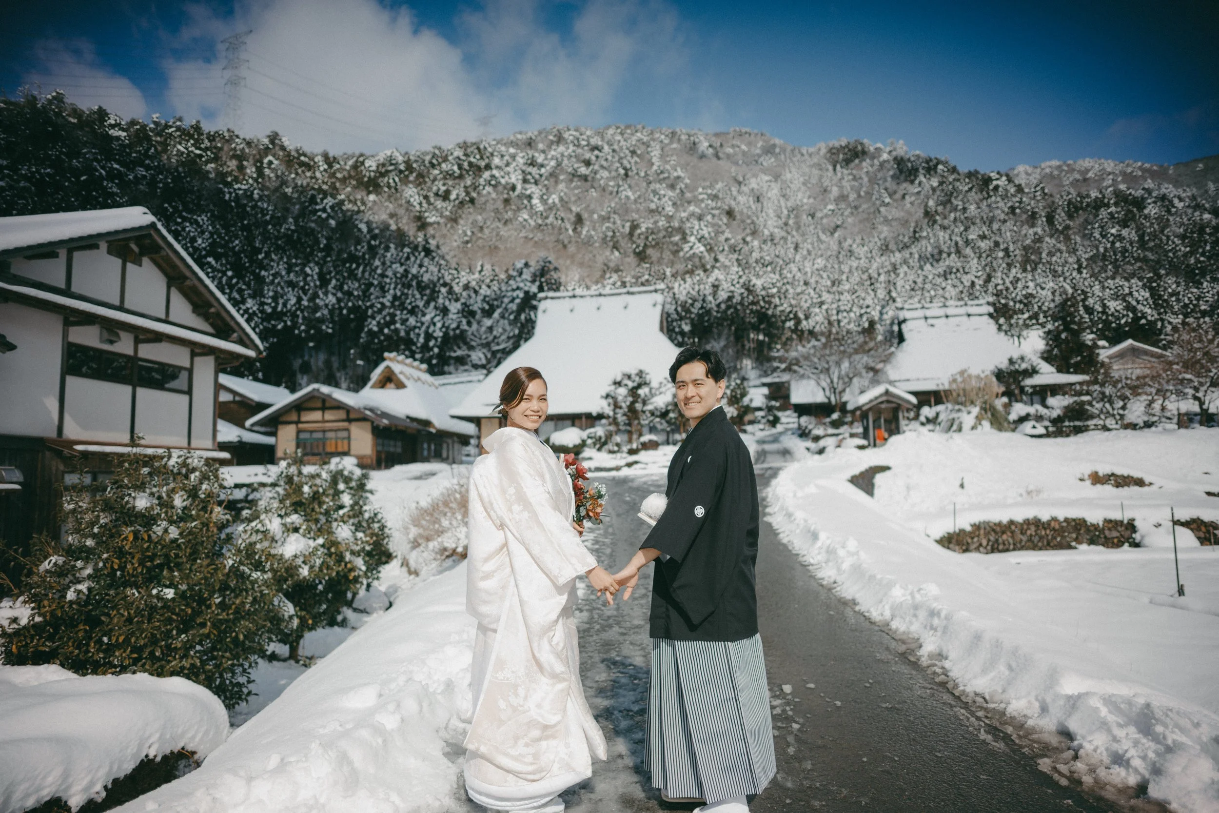 A newly married couple dressed in traditional Japanese wedding attire holding hands and smiling on a snowy street in a rural village, with snow-covered houses and mountains in the background.