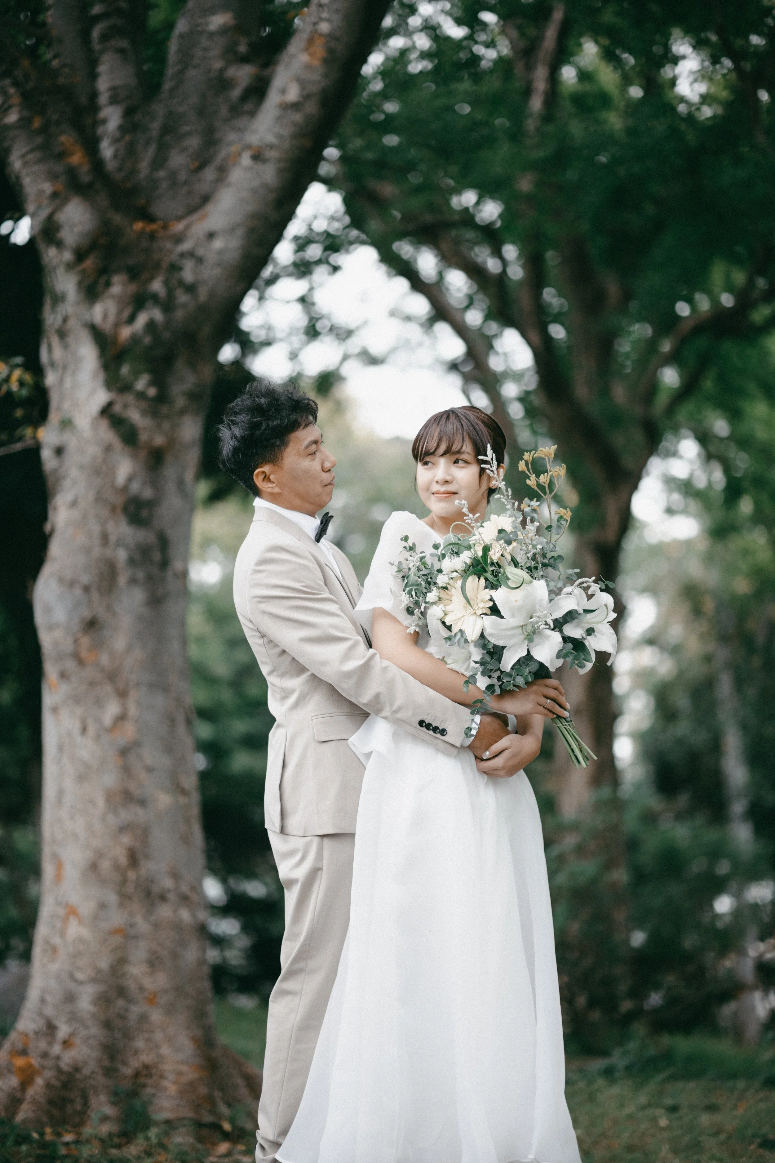 A bride and groom in wedding attire standing outdoors under a large tree, with the groom holding the bride and she holding a large bouquet of white flowers and greenery.