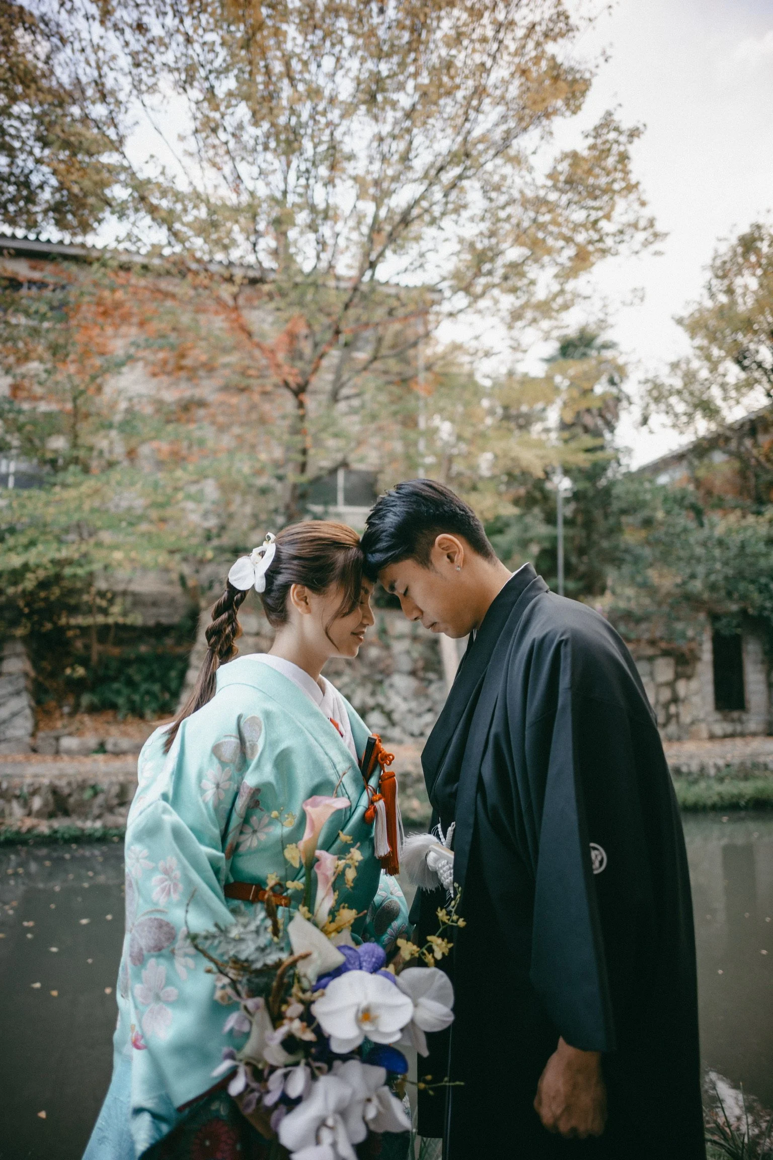 A young Japanese couple dressed in traditional wedding attire, standing close with foreheads touching, holding a bouquet of flowers outdoors by a pond with autumn trees in the background.