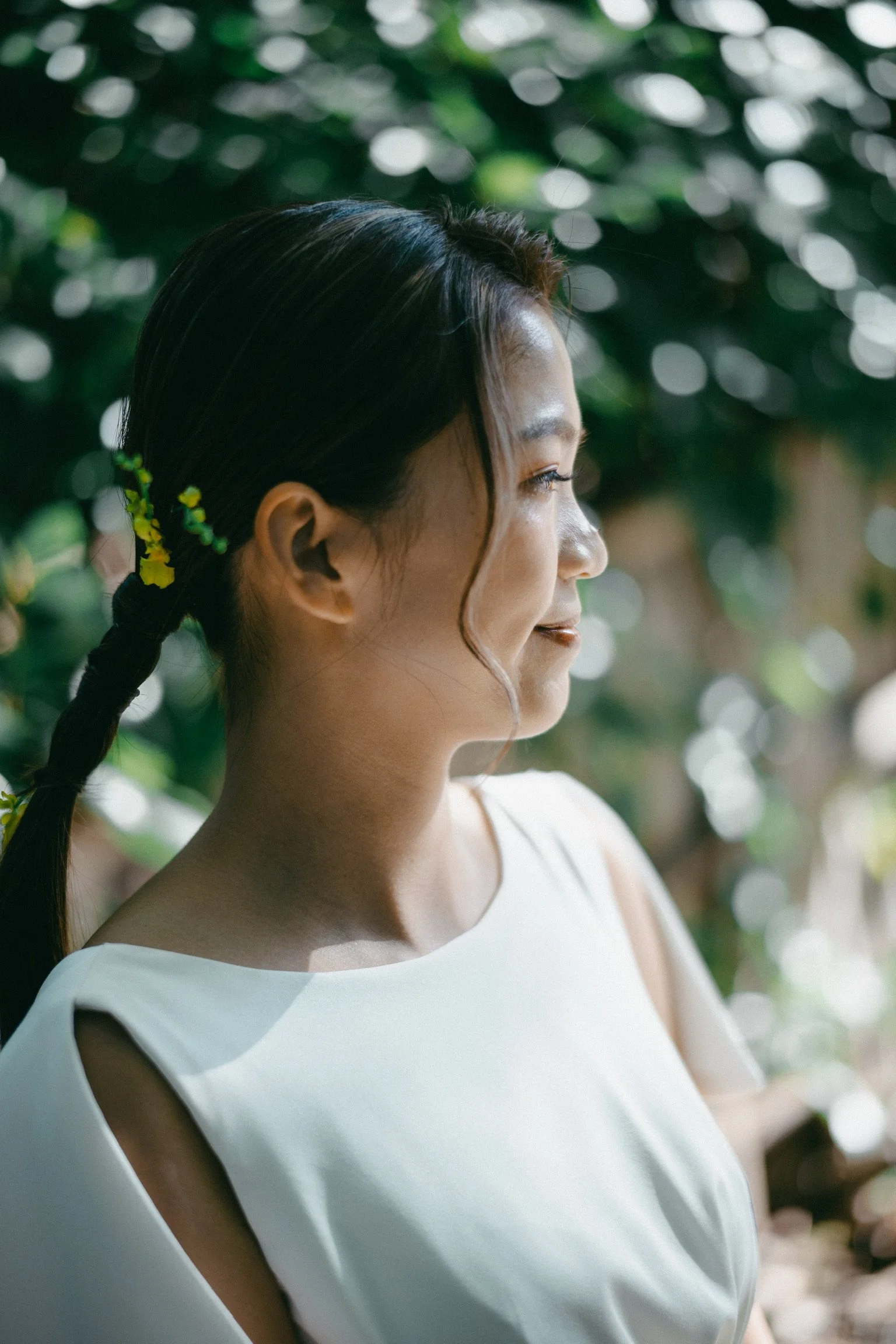 Profile of a young woman with dark hair in a braid and a gentle smile, outdoors with green foliage in the background.