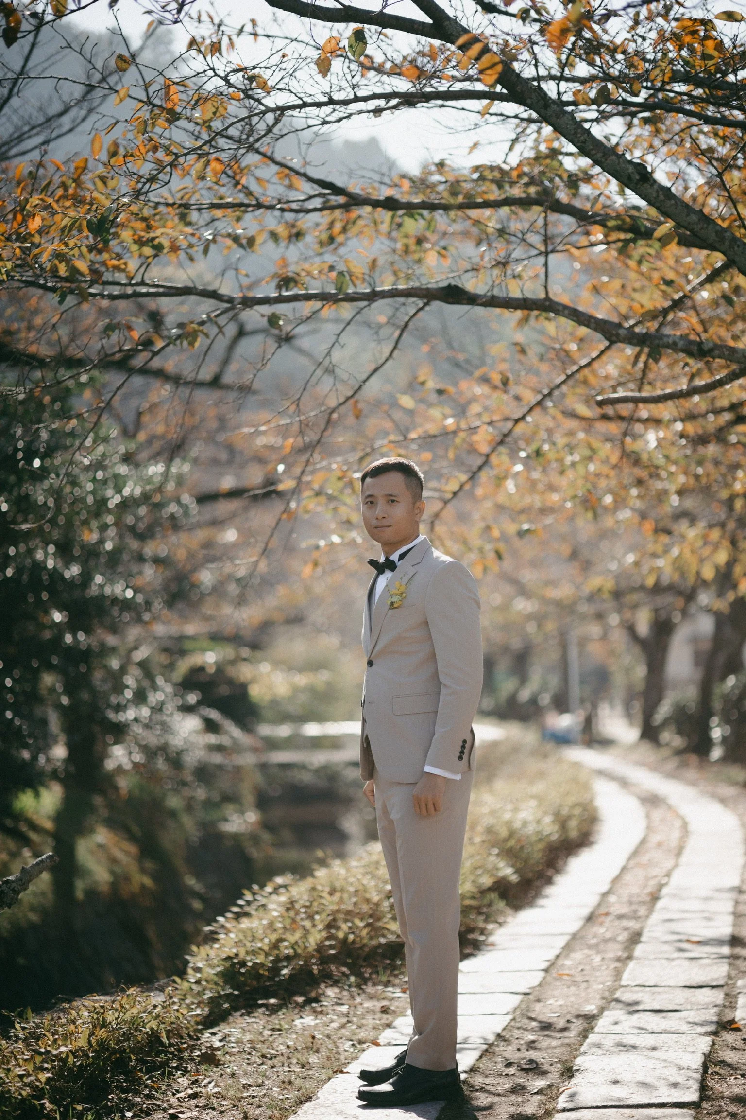 A man dressed in a beige tuxedo with a black bow tie, standing on a curved stone path in a park with fall-colored trees.
