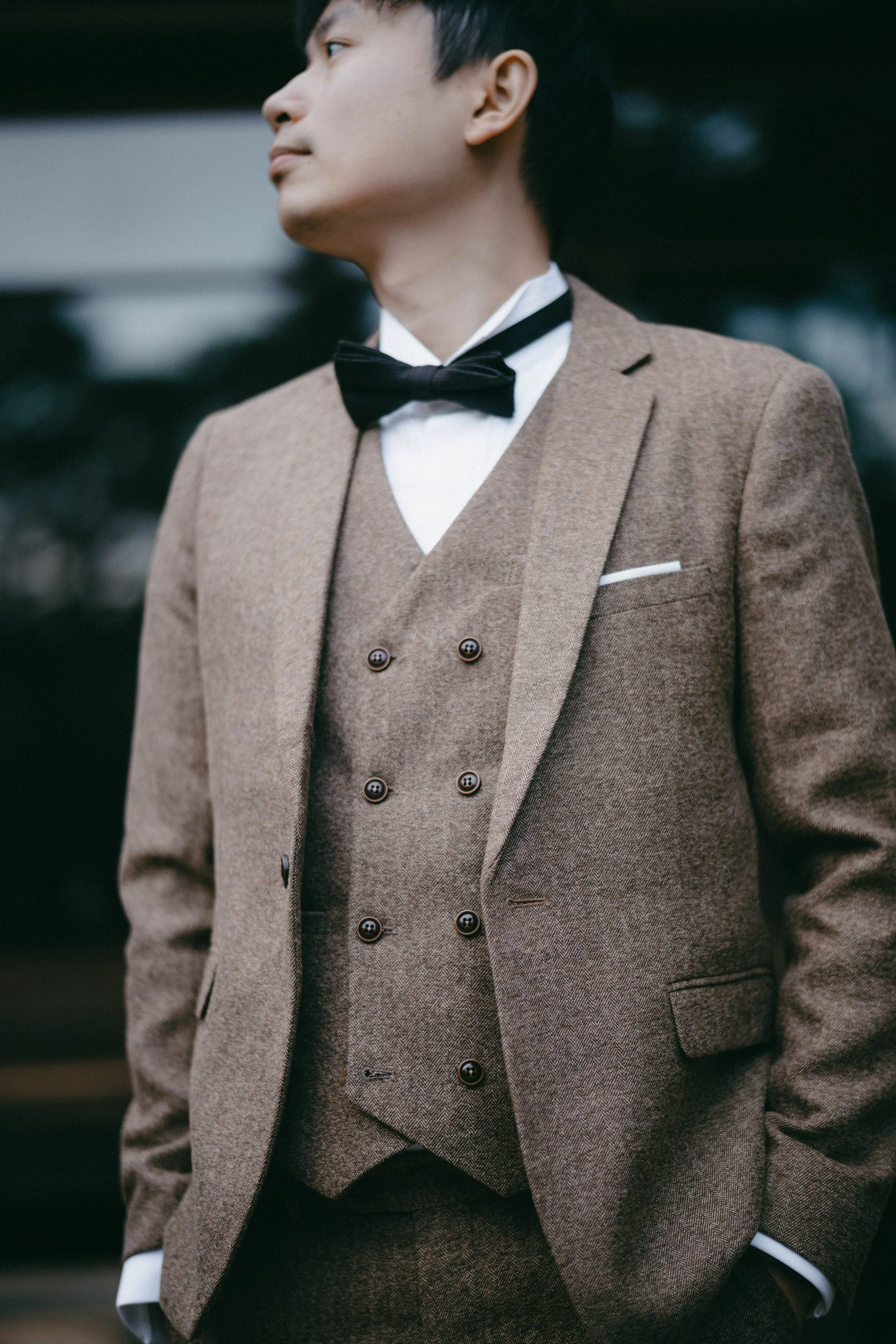 A man in a brown suit with a black bow tie, white shirt, and tan vest, standing outdoors with a contemplative expression.