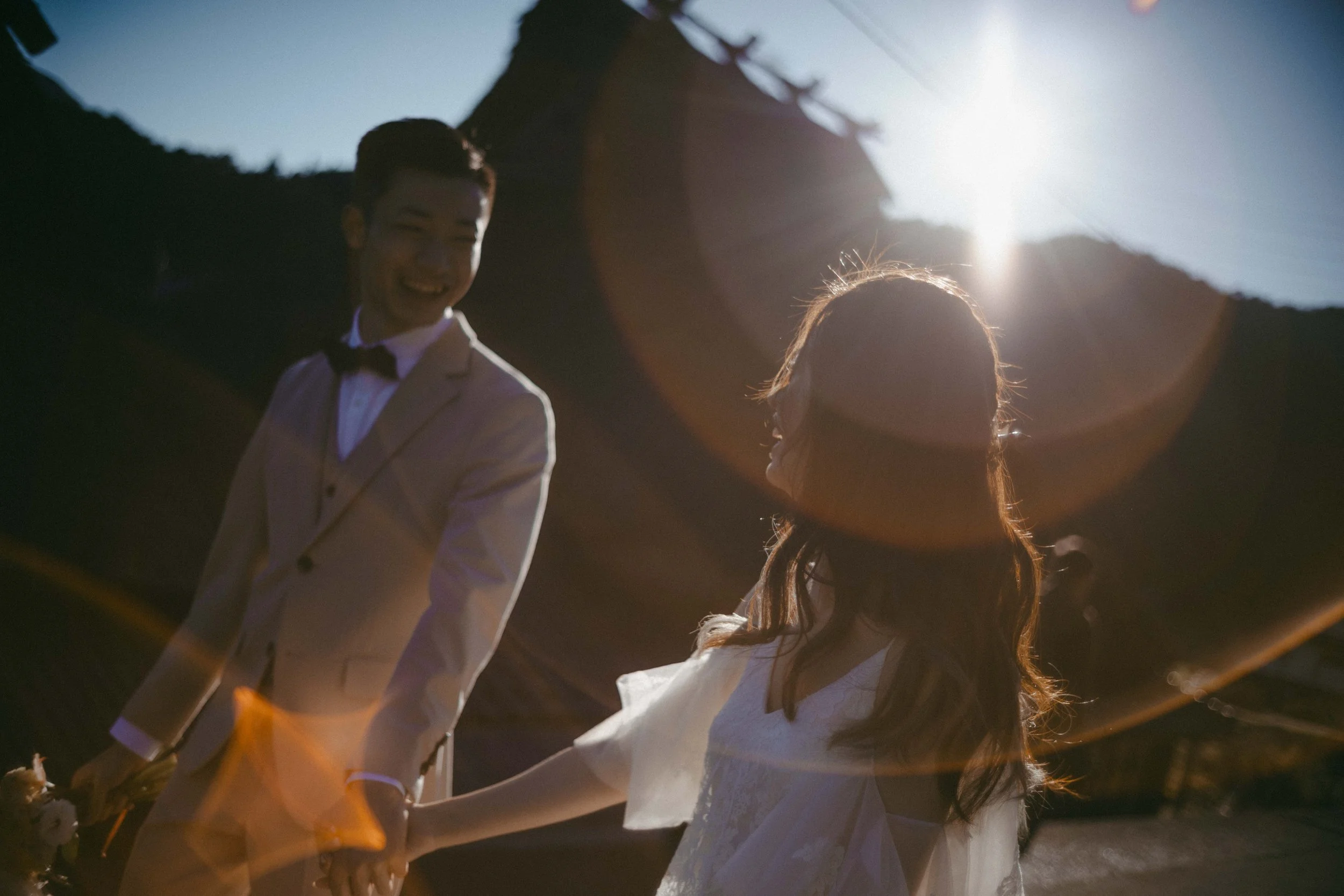A couple holding hands outdoors during sunset, with the man in a white suit with a black bow tie and the woman in a white dress, smiling at each other.