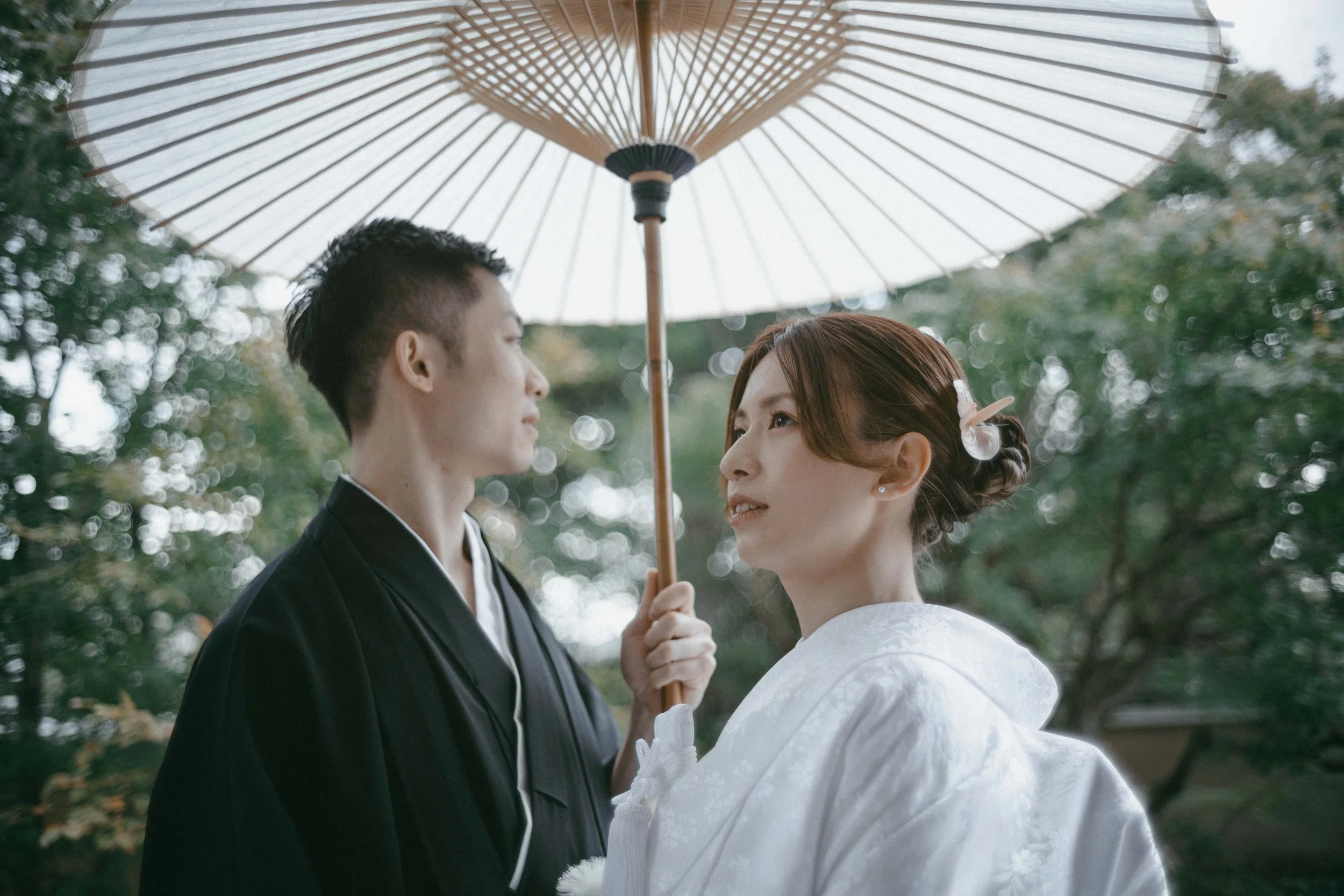A couple dressed in traditional Japanese wedding attire standing outdoors under a parasol, gazing at each other with trees in the background.