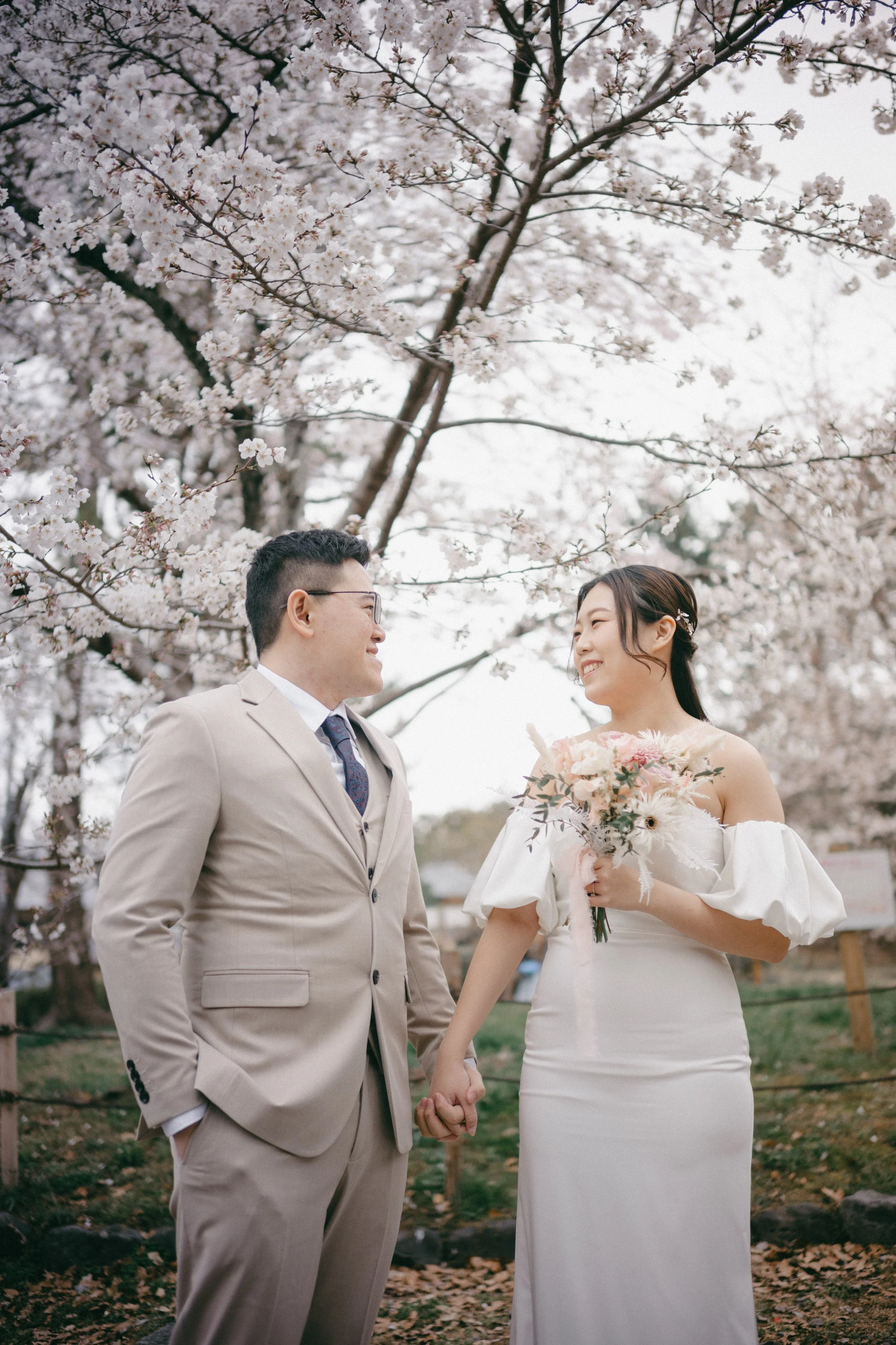 A bride and groom holding hands and smiling at each other outdoors under blooming cherry blossom trees.