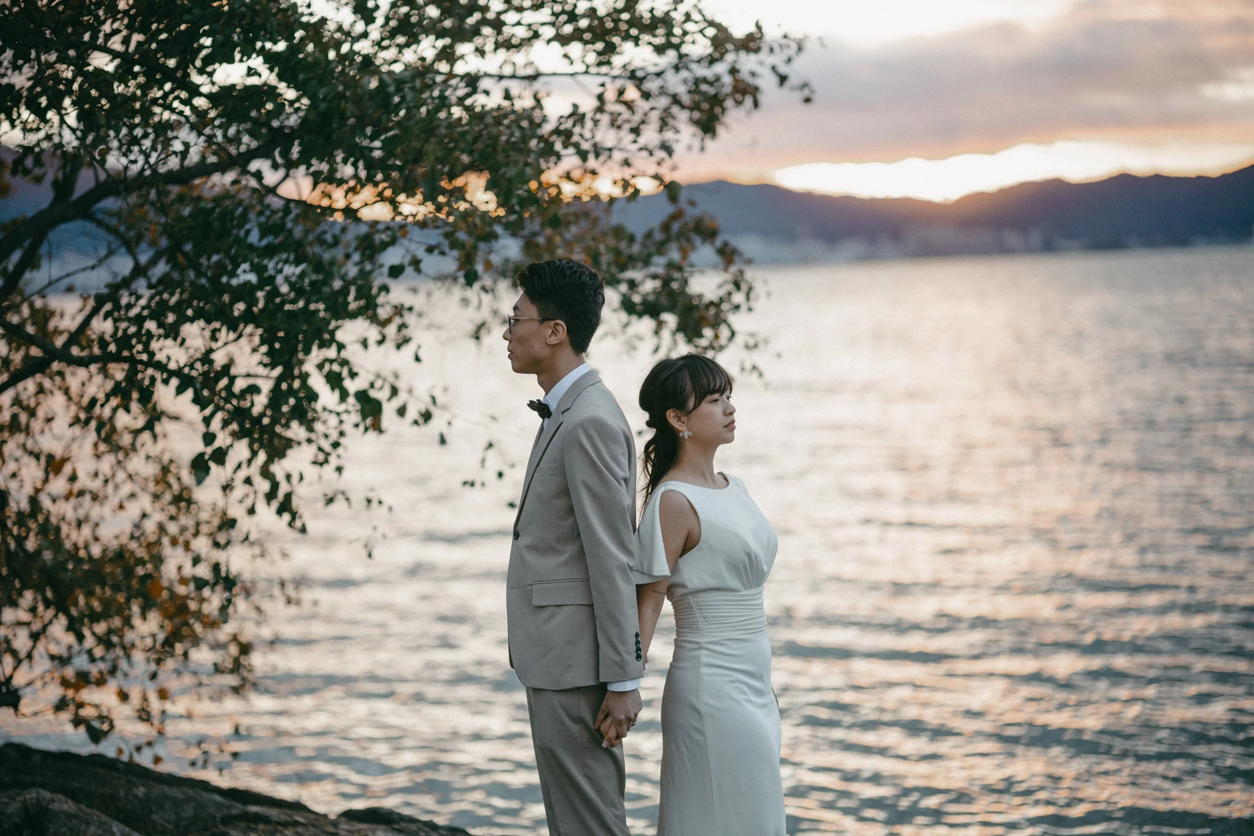 A couple dressed in wedding attire stands back to back near a body of water during sunset. The man wears a light-colored suit and glasses, and the woman wears a white dress with cold shoulders. There are trees and mountains in the background.